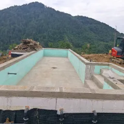 Fundament basenu z izolacją termiczną i hydroizolacją, na tle góry. Widoczny mały, czerwony koparka i stos drewna. Etap budowy basenu.