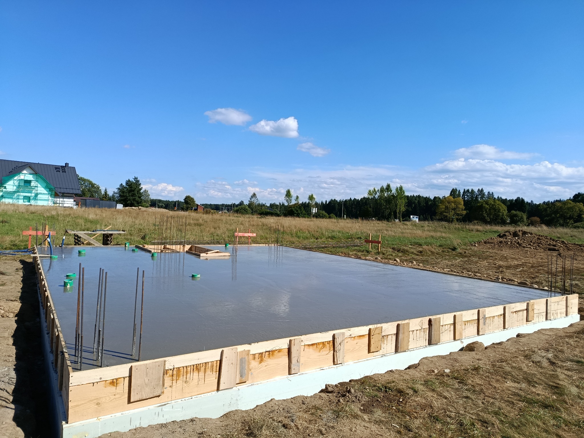 Zalany fundament pod budowę domu jednorodzinnego, szalunek z desek, wystające pręty zbrojeniowe, widok na okolicę w słoneczny dzień.
