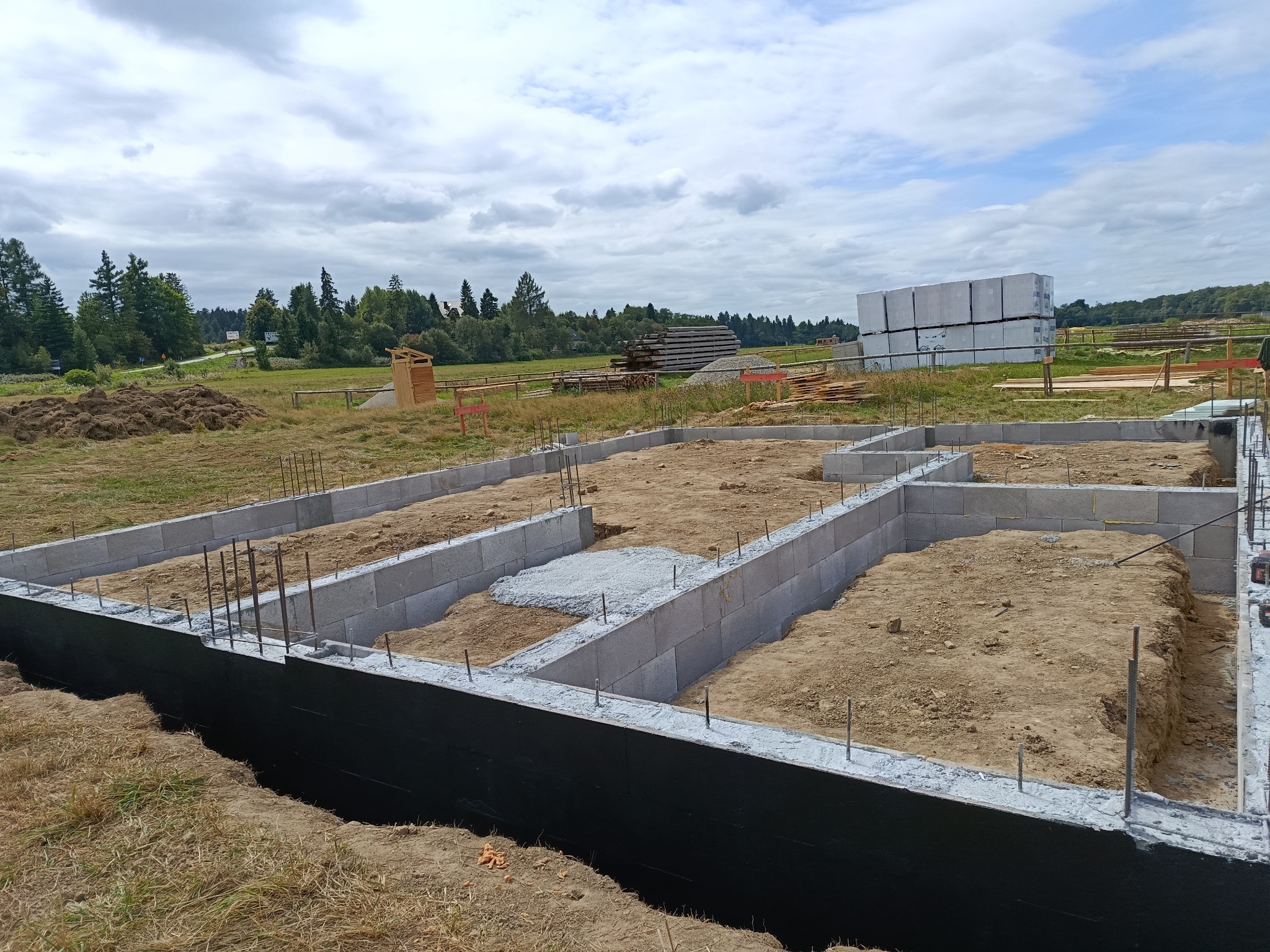 Zabezpieczone przed wilgocią fundamenty z betonu komórkowego, częściowo wypełnione piaskiem, z wystającymi prętami zbrojeniowymi. W tle składowane bloczki.