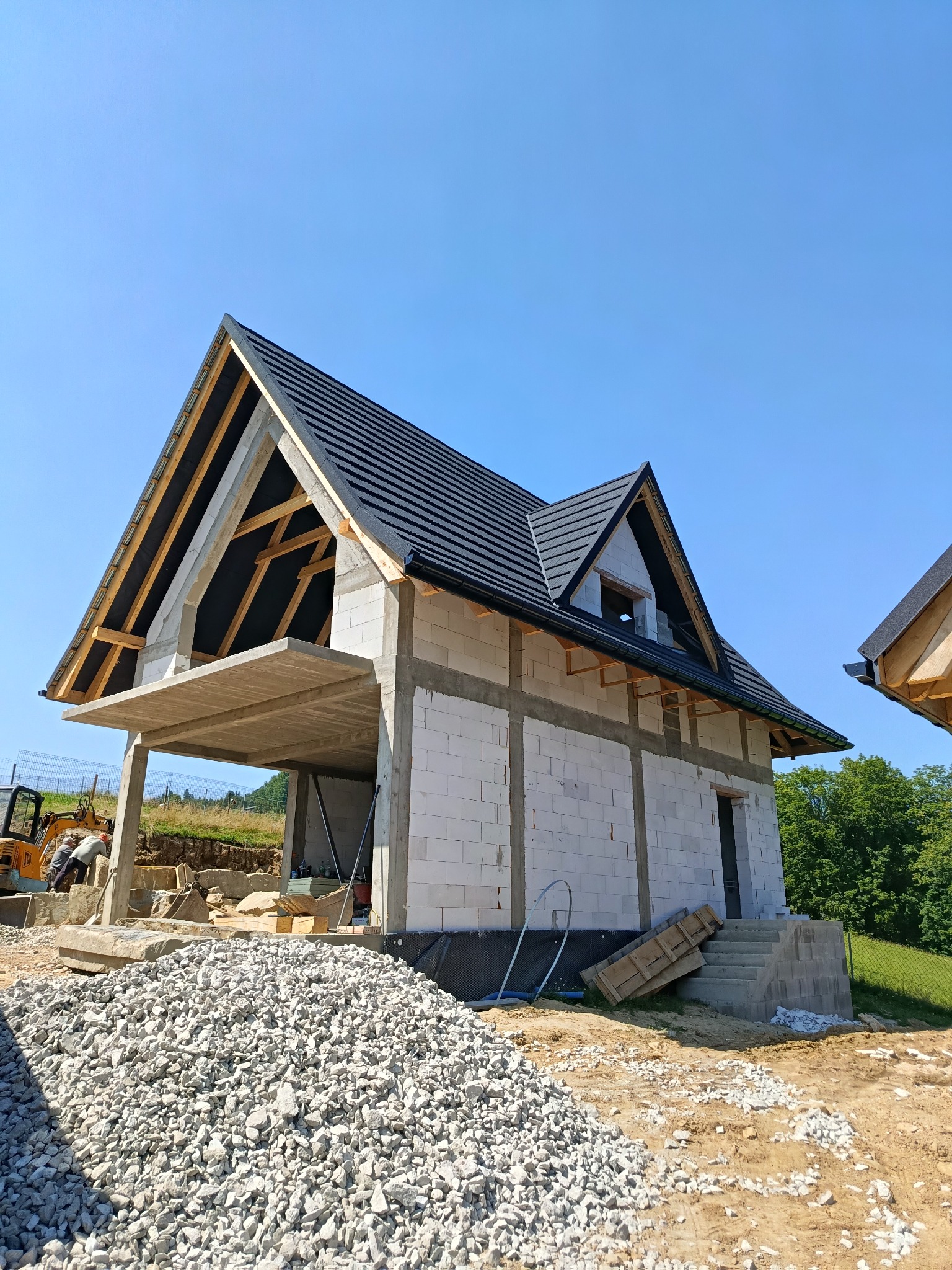 Wznoszenie domu z białych bloczków: widoczna konstrukcja dachu z czarną dachówką, betonowy strop nad garażem, wzmocnienia pionowe. Na pierwszym planie hałda gruzu.