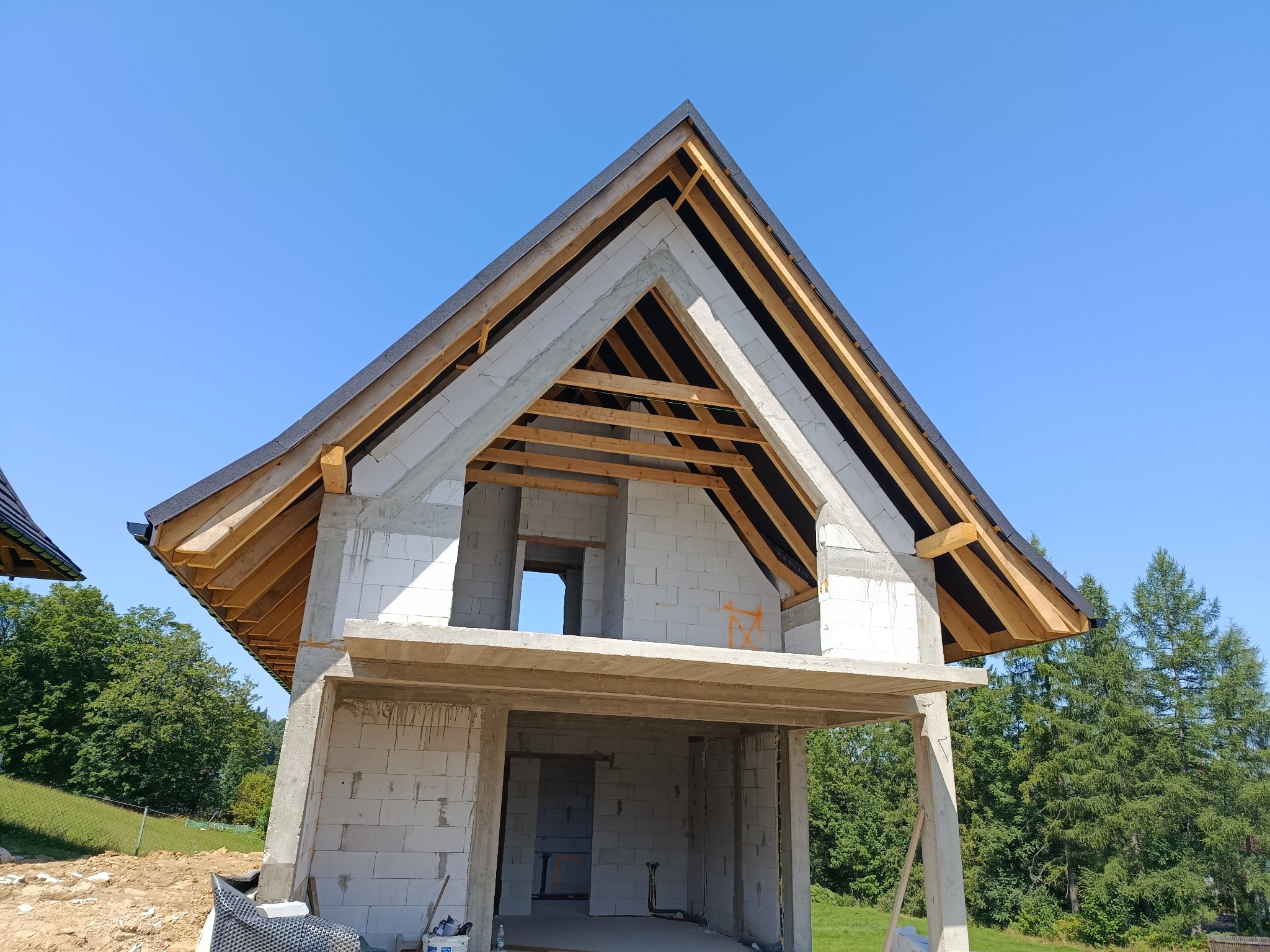 Surowy stan domu z białych bloczków, z widoczną konstrukcją dachu i drewnianymi belkami, na tle błękitnego nieba i zieleni drzew. Widok z dołu.
