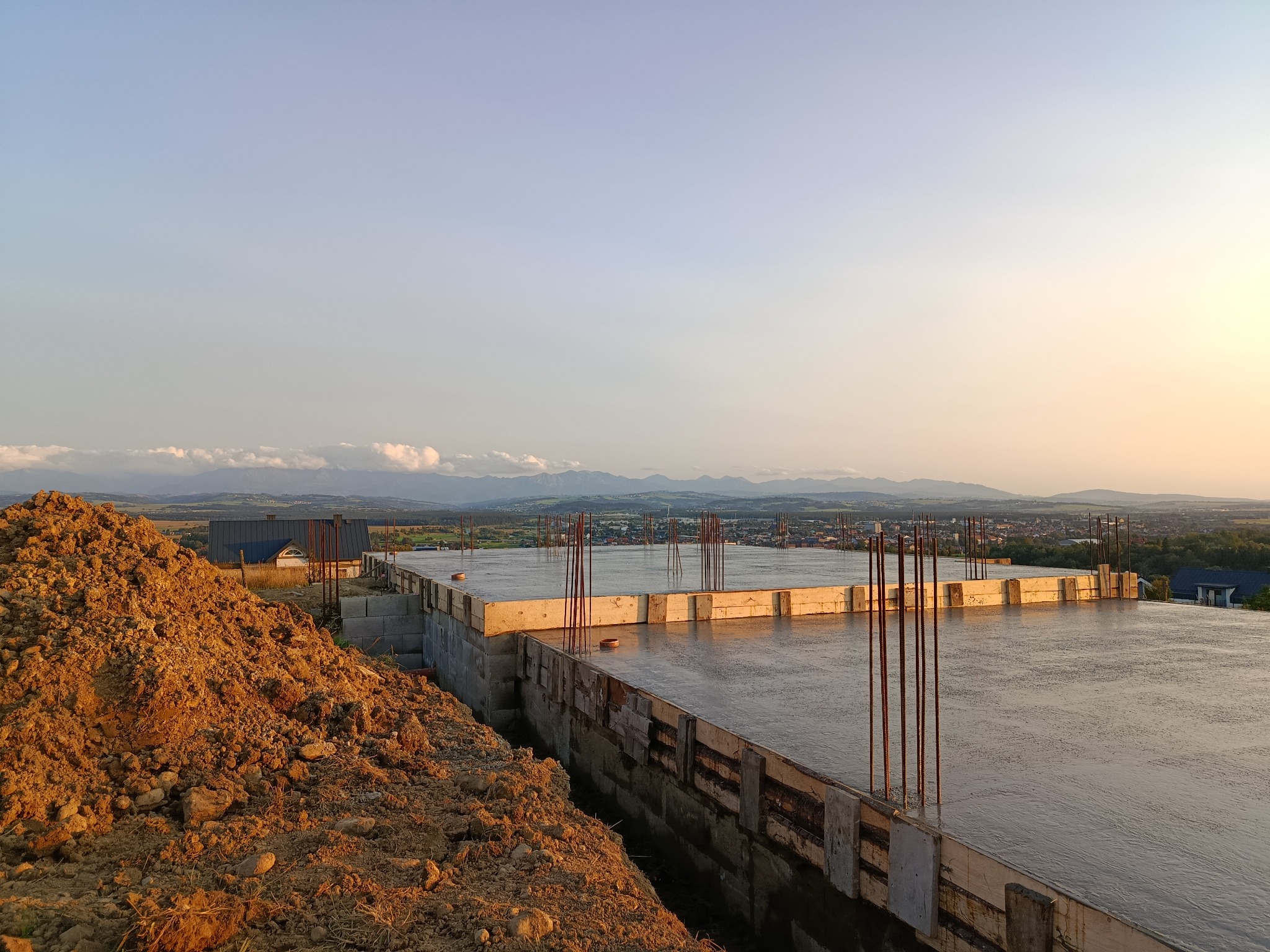 Zalany fundament płyty budynku w trakcie budowy, z wystającymi prętami zbrojeniowymi. W tle widok na panoramę miasta i góry w oddali, oświetlone zachodzącym słońcem.