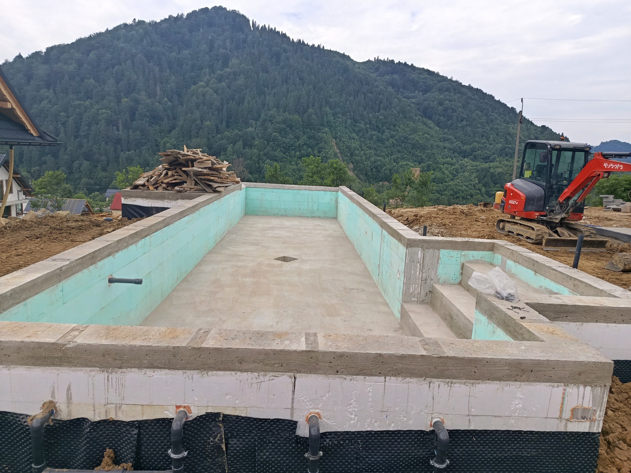 Fundament basenu z izolacją termiczną i hydroizolacją, na tle góry. Widoczny mały, czerwony koparka i stos drewna. Etap budowy basenu.