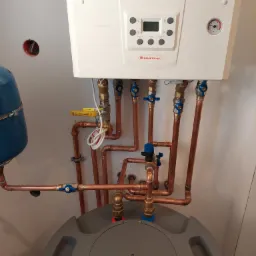 Biały piec gazowy marki Immergas, model Victrix Tera, z podłączeniami z miedzianych rur i zaworami, zamontowany na ścianie, z widocznym zbiornikiem wyrównawczym i szarą pokrywą zasobnika ciepłej...