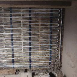 Rury ogrzewania podłogowego przymocowane do trzciny na ścianie w trakcie instalacji, widoczne mocowania i fragment ściany z gliny.