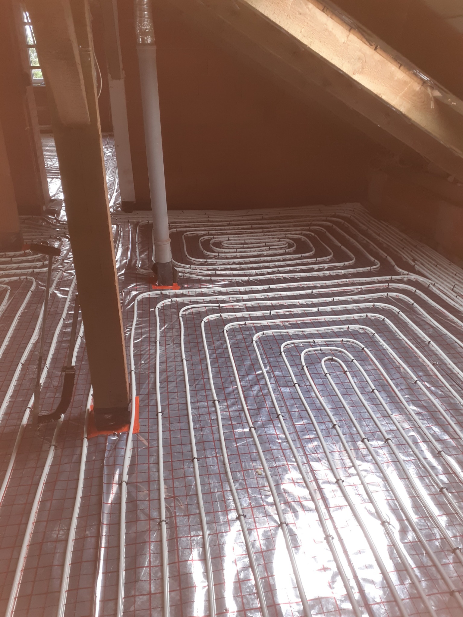 Instalacja ogrzewania podłogowego w poddaszu: białe rury grzewcze ułożone w spiralny wzór na srebrnej folii izolacyjnej z czerwoną siatką, widoczne elementy konstrukcji drewnianej dachu i pionowe...