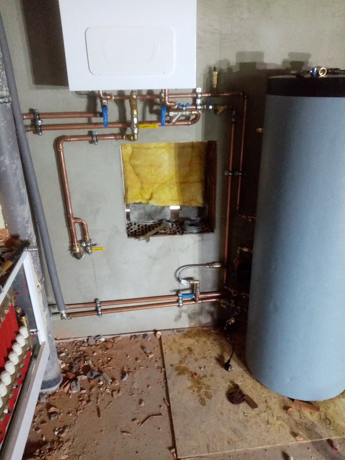 Świeżo zamontowany piec gazowy z podgrzewaczem wody, widoczne miedziane rury z zaworami, izolacja ściany, narzędzia w niszy i bałagan na podłodze.