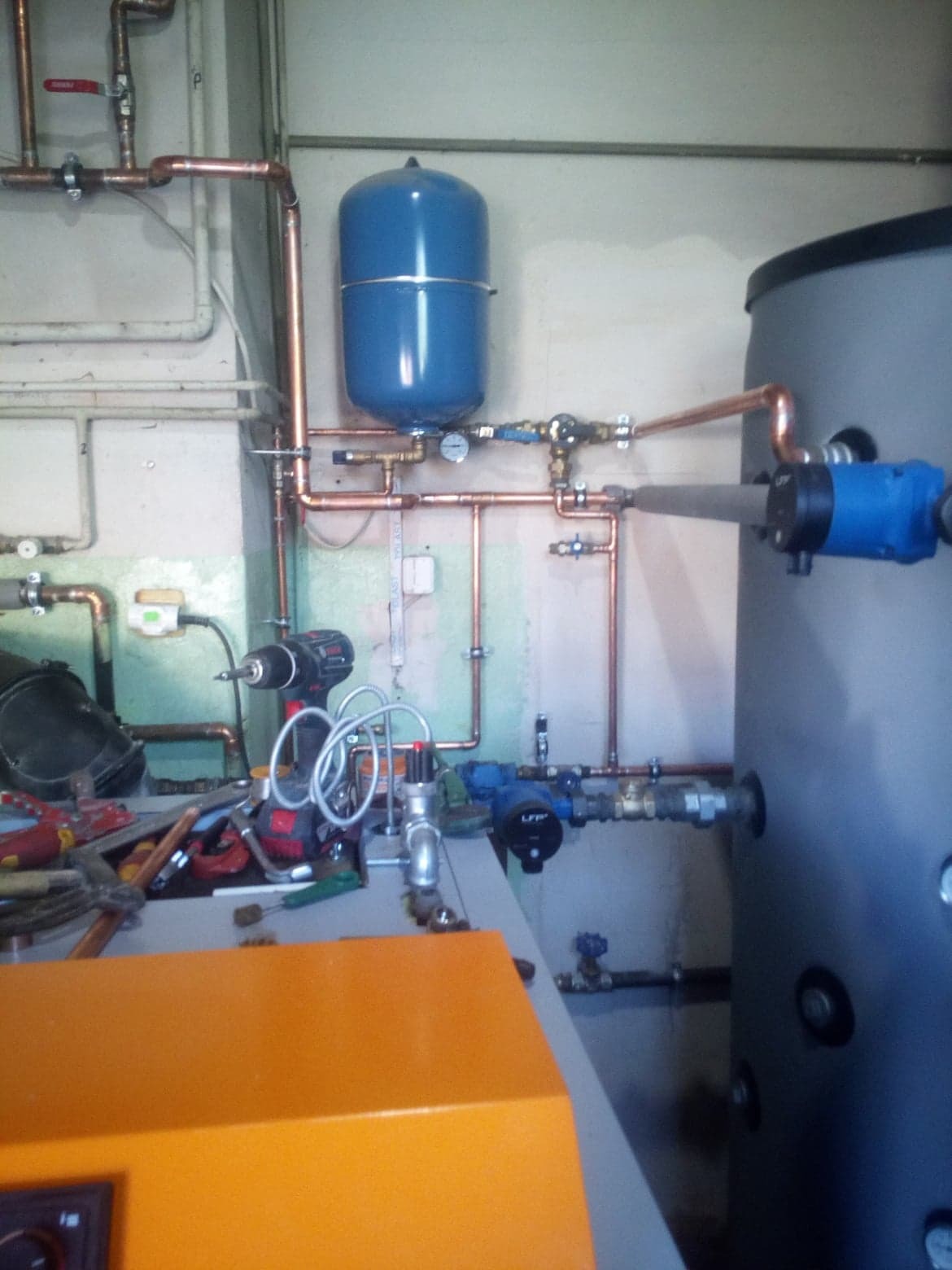 Instalacja grzewcza z miedzianymi rurami, zbiornikiem wyrównawczym i pompą obiegową, widoczne narzędzia na blacie.
