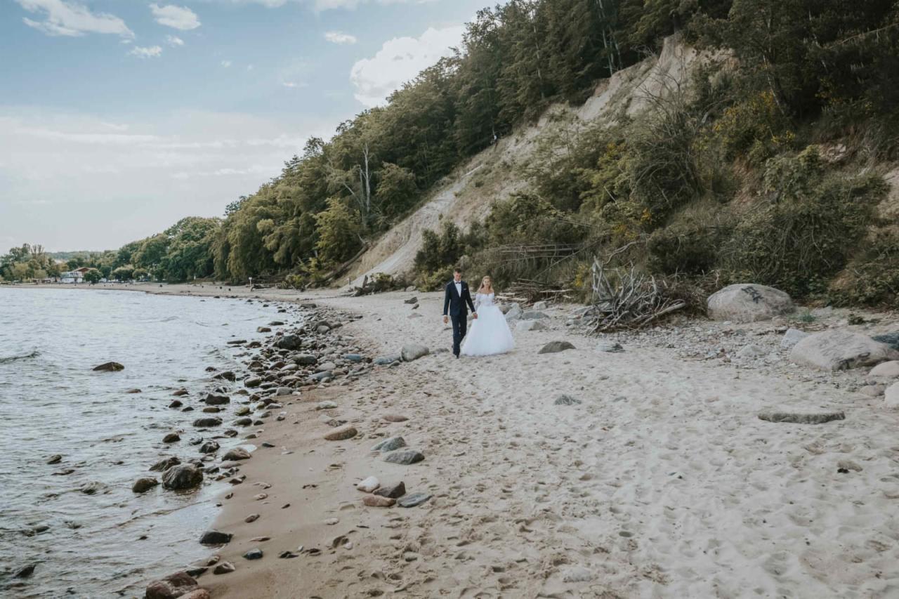 Para młoda spaceruje po piaszczystej plaży wzdłuż morza, zalesione wzgórze w tle, panna młoda w białej sukni z długim rękawem, pan młody w granatowym garniturze.