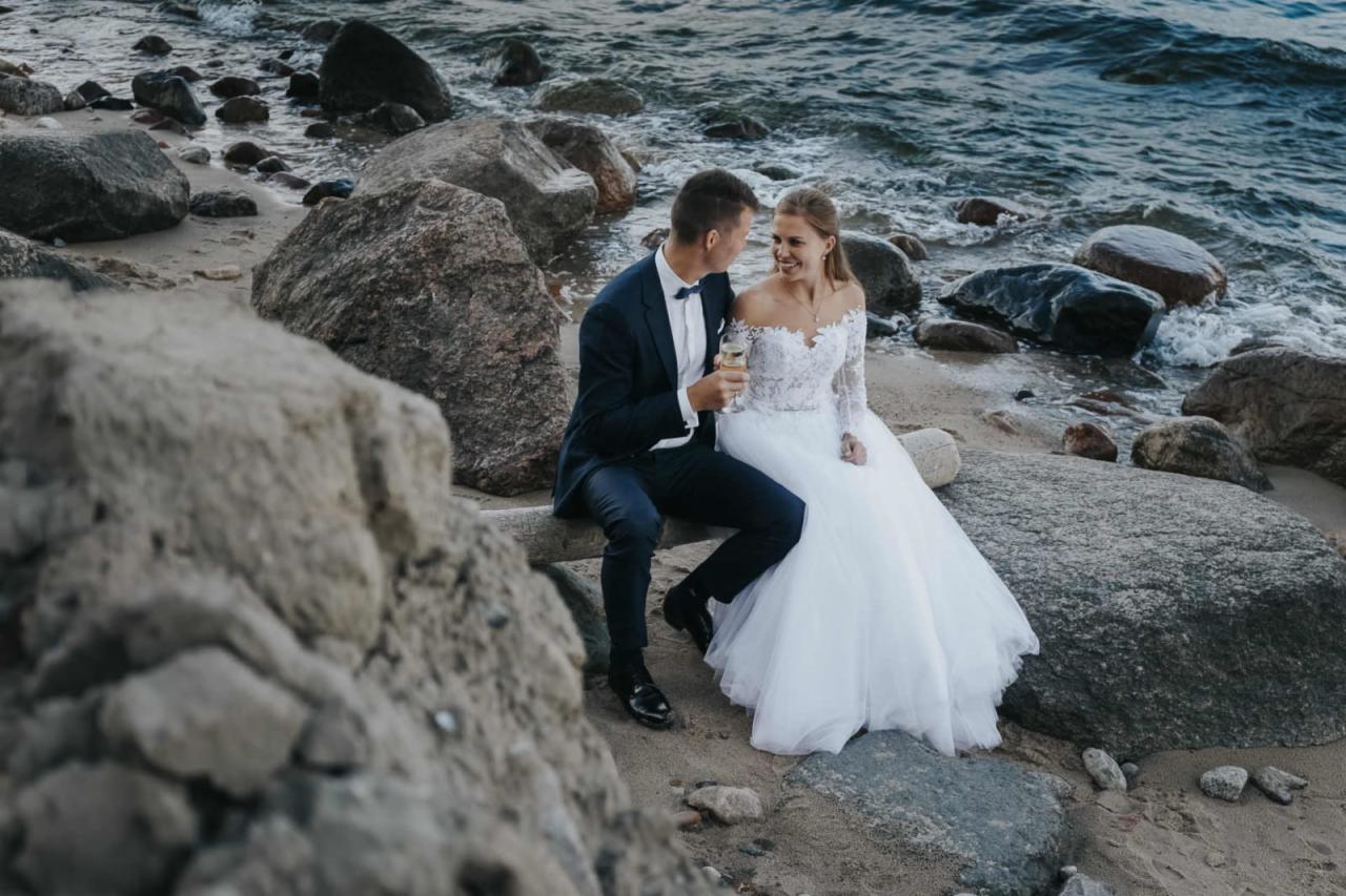 Para młoda siedzi na pniu drzewa na plaży z kamieniami, panna młoda w koronkowej sukni z długim rękawem, pan młody w granatowym garniturze trzyma kieliszek szampana, patrzą na siebie z uśmiechem,...