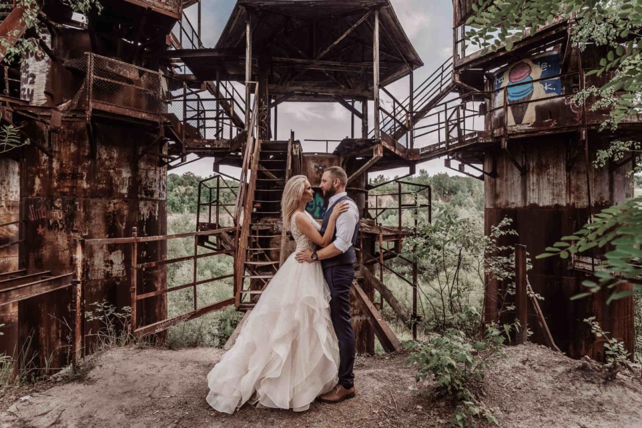 Para młoda w sukni ślubnej i garniturze pozuje na tle opuszczonej, zardzewiałej konstrukcji przemysłowej z graffiti i zielenią w tle.