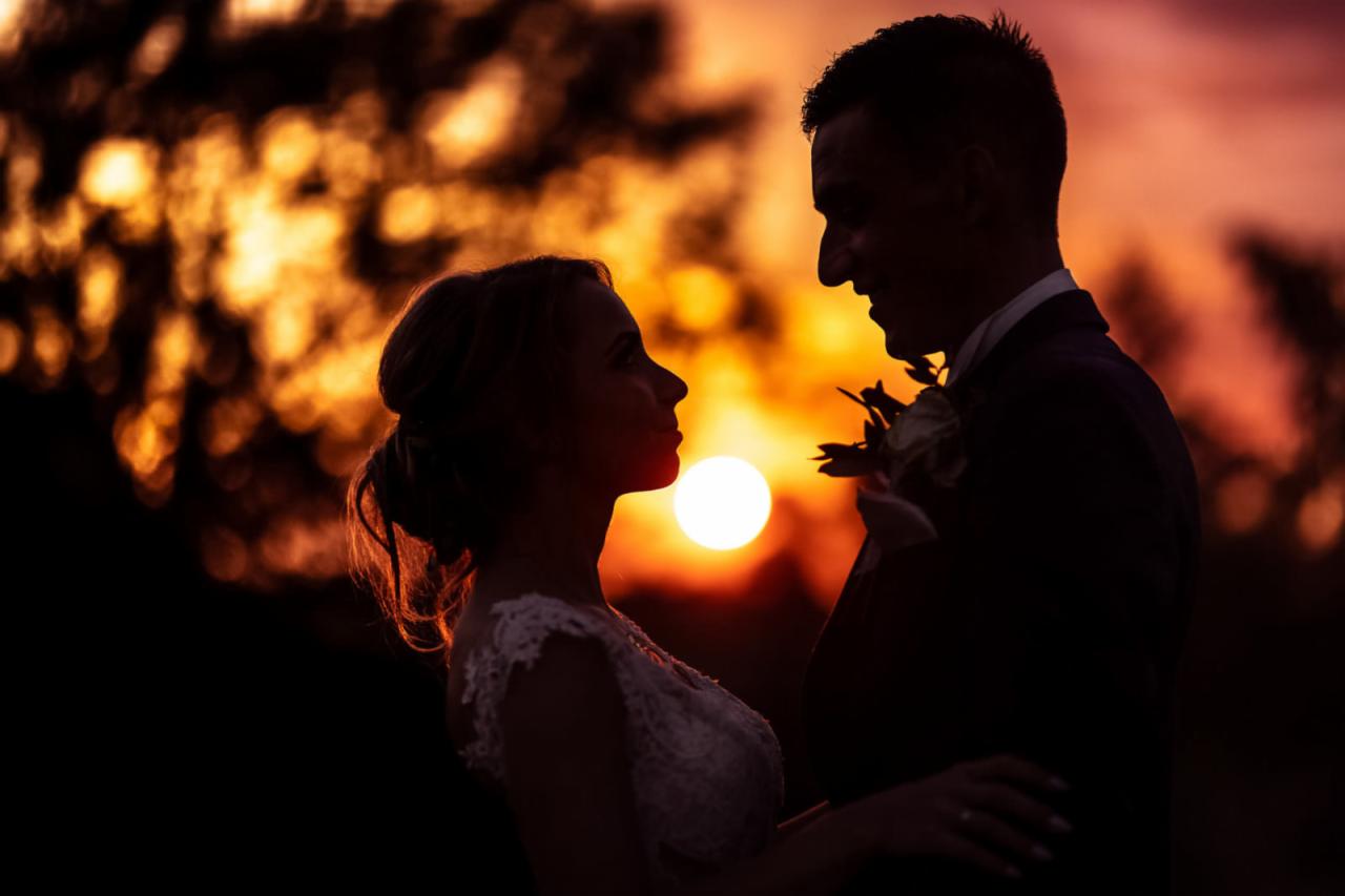 Sylwetki pary młodej na tle zachodzącego słońca, ona w koronkowej sukni, on w garniturze z butonierką, oboje patrzą na siebie z uśmiechem.