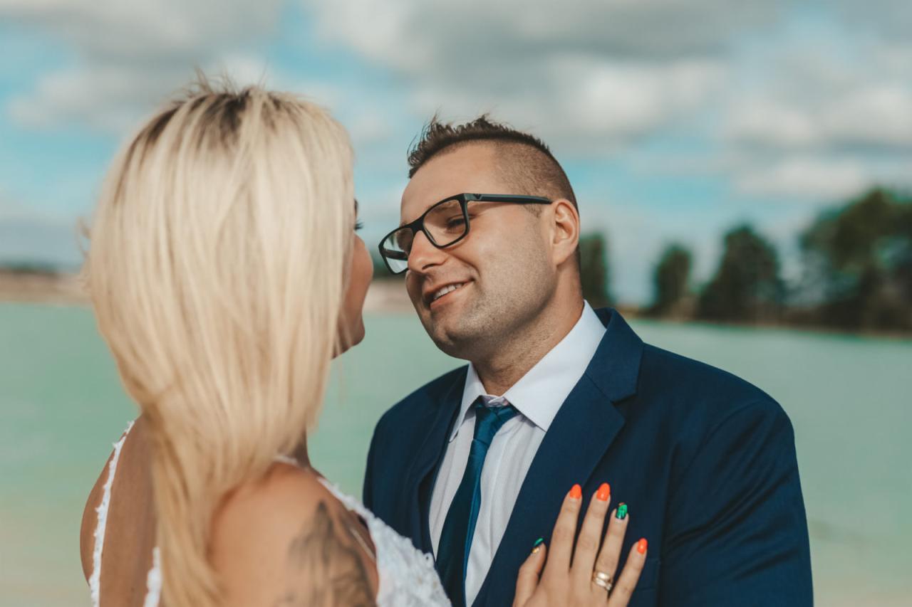 Para młoda wpatrzona w siebie na tle turkusowej wody i błękitnego nieba z chmurami; panna młoda z blond włosami i tatuażem na plecach, pan młody w okularach i granatowym garniturze z niebieskim...