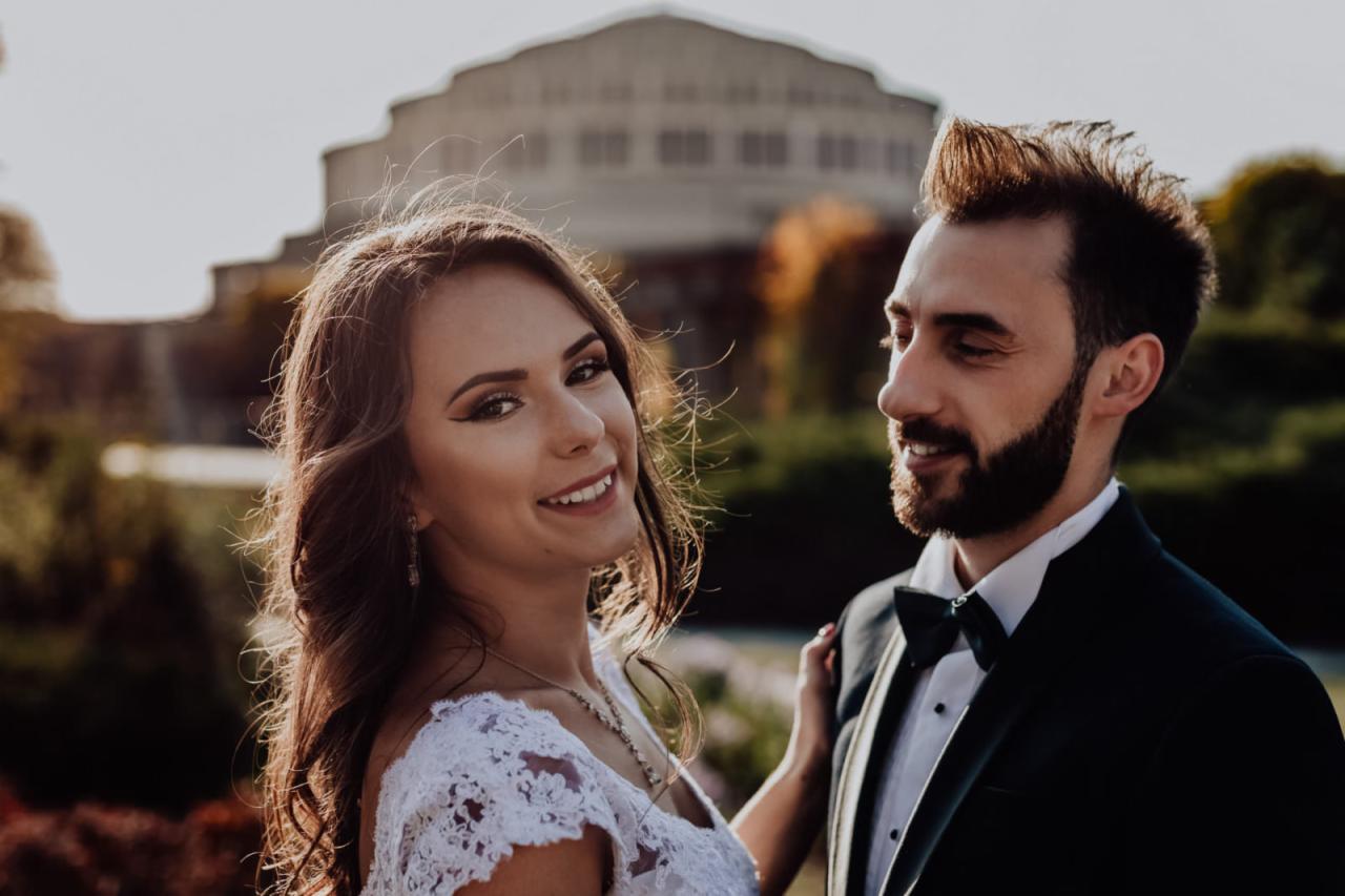 Uśmiechnięta panna młoda w koronkowej sukni i pan młody w smokingu z muszką, na tle Hali Stulecia we Wrocławiu w ciepłym świetle zachodzącego słońca.