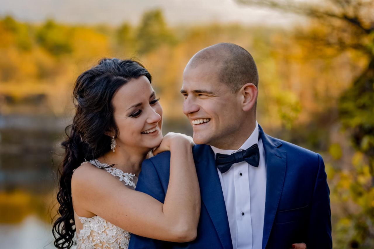 Uśmiechnięta panna młoda w koronkowej sukni opiera głowę na ramieniu pana młodego w granatowym garniturze z muchą, na tle jesiennego krajobrazu z jeziorem i drzewami.