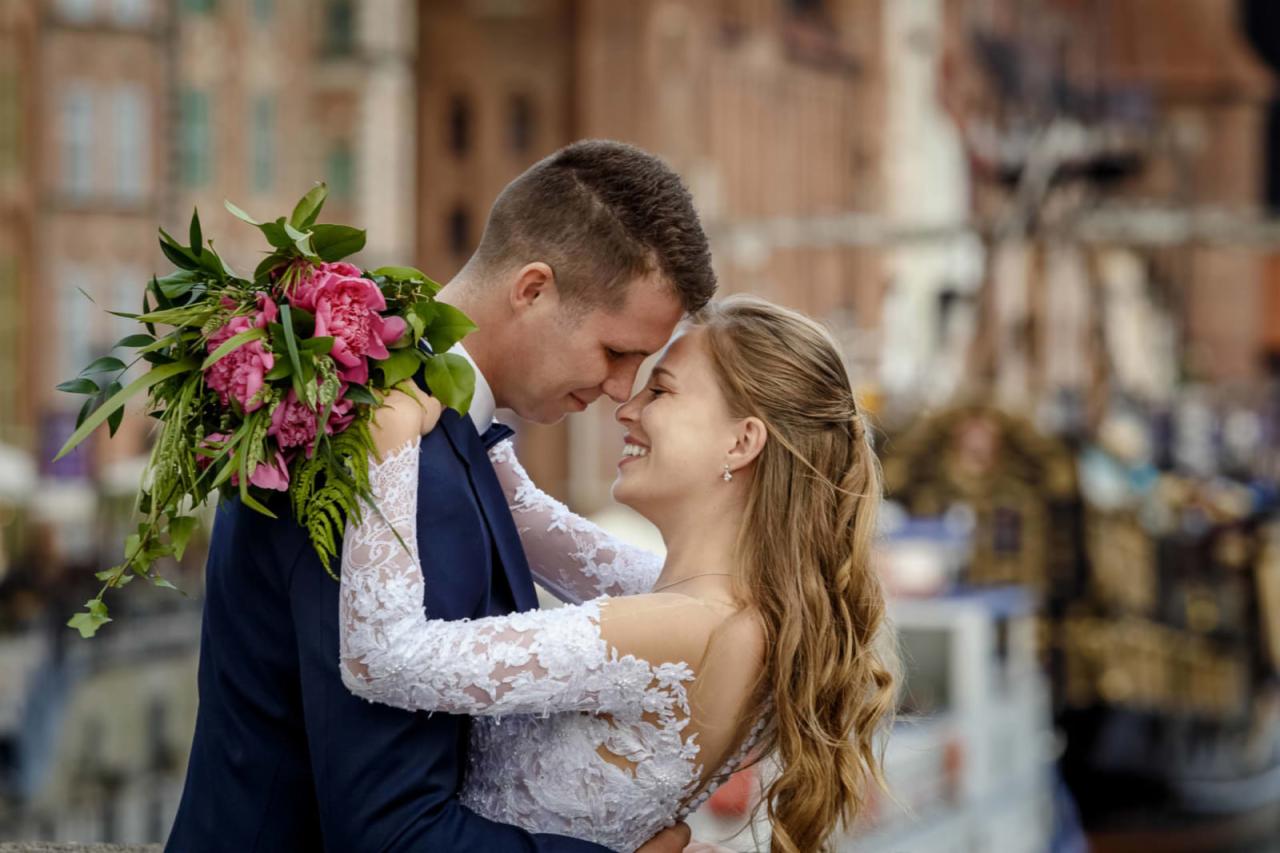 Uśmiechnięta para młoda, panna młoda w sukni z koronkowymi rękawami trzyma bukiet różowych piwonii, dotykają się czołami na tle miejskiej zabudowy.
