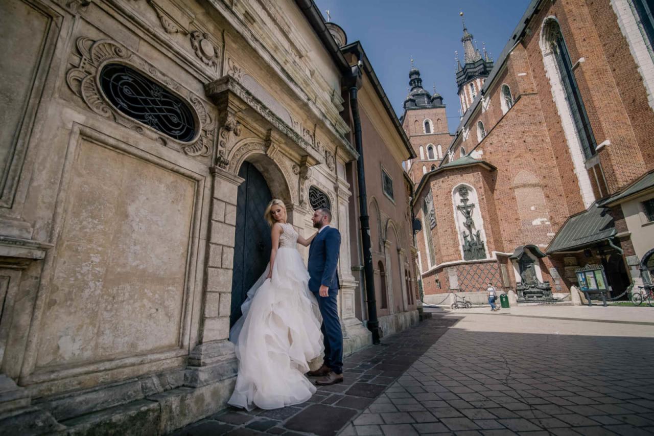Para młoda pozuje przy zabytkowej kamienicy z kościołem w tle. Panna młoda w białej sukni z falbanami, pan młody w granatowym garniturze.