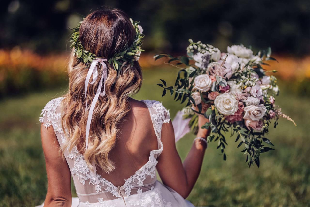 Panna młoda z tyłu, w koronkowej sukni ślubnej, z wiankiem na głowie i bukietem w ręku, siedząca na trawie.