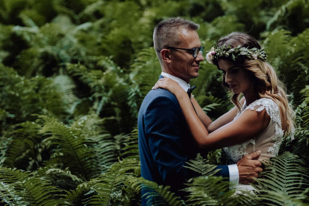 Para młoda w objęciach, on w granatowym garniturze i okularach, ona w białej sukni z wiankiem na głowie, otoczeni gęstą zielenią paproci.