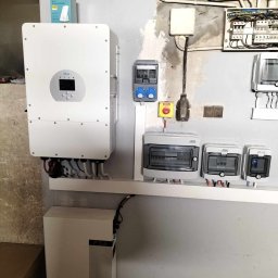 BAHAUS TOMASZ BACH - Instalacja magazynu energii: biały inwerter Deye, bateria pod nim, skrzynki bezpiecznikowe i rozdzielcze na ścianie, widoczny router TP-Link