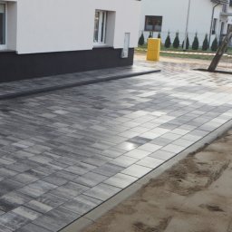 BAHAUS TOMASZ BACH - Ułożona, szara kostka brukowa na podjeździe przed nowoczesnym domem z białymi ścianami i ciemnoszarym cokołem. Widoczne obrzeża i fragment trawnika w tle.