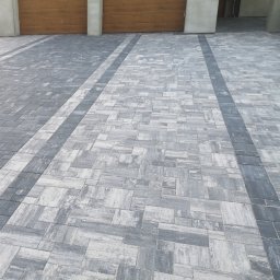 BAHAUS TOMASZ BACH - Ułożona kostka brukowa w odcieniach szarości prowadząca do garażu z drewnianymi drzwiami. Widok z perspektywy niskiego kąta.