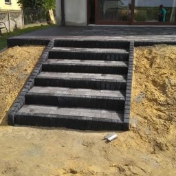 BAHAUS TOMASZ BACH - Schody z kostki brukowej prowadzące do domu z dużym oknem tarasowym. Widoczne niedokończone prace ziemne wokół schodów. Użyto ciemnoszarej kostki.