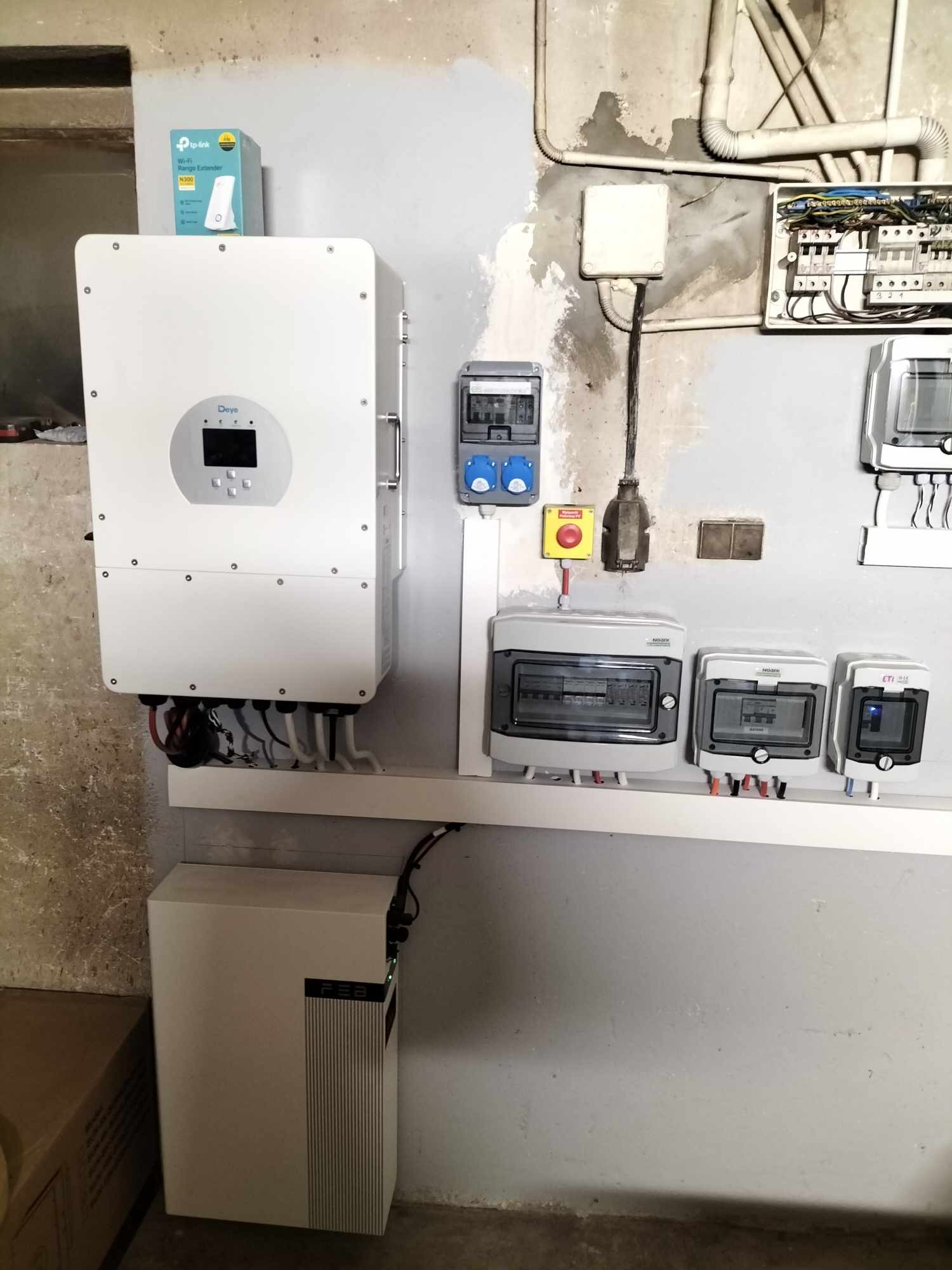 Instalacja magazynu energii: biały inwerter Deye, bateria pod nim, skrzynki bezpiecznikowe i rozdzielcze na ścianie, widoczny router TP-Link