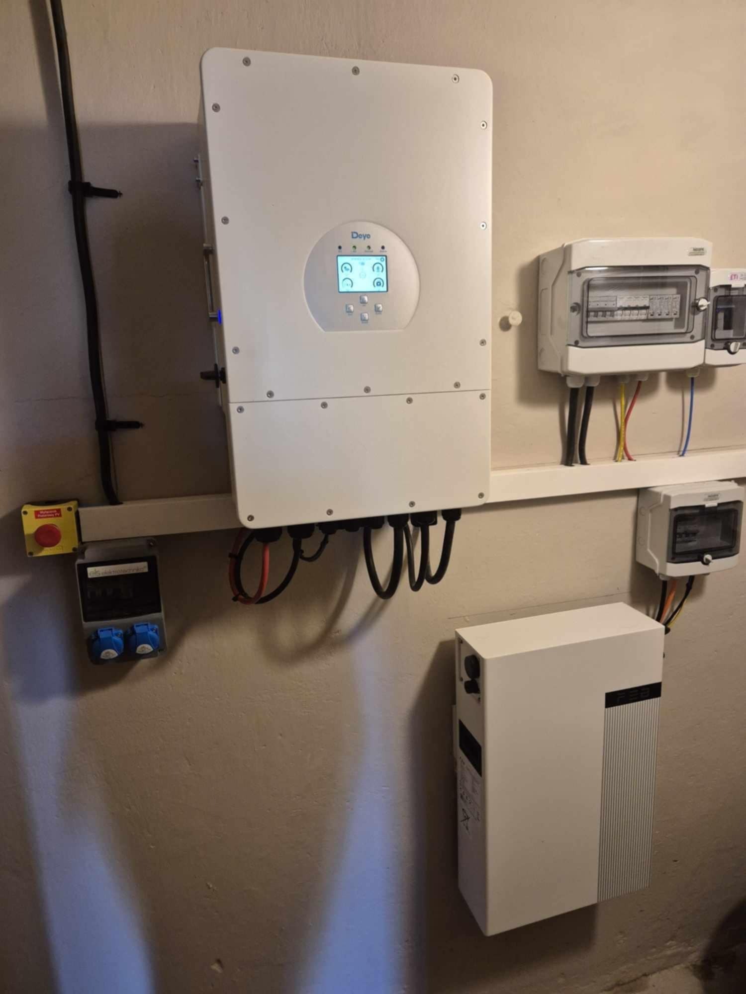 Biały inwerter fotowoltaiczny marki Deye z wyświetlaczem, skrzynka bezpiecznikowa, licznik energii elektrycznej i akumulator energii zamontowane na ścianie w pomieszczeniu technicznym.
