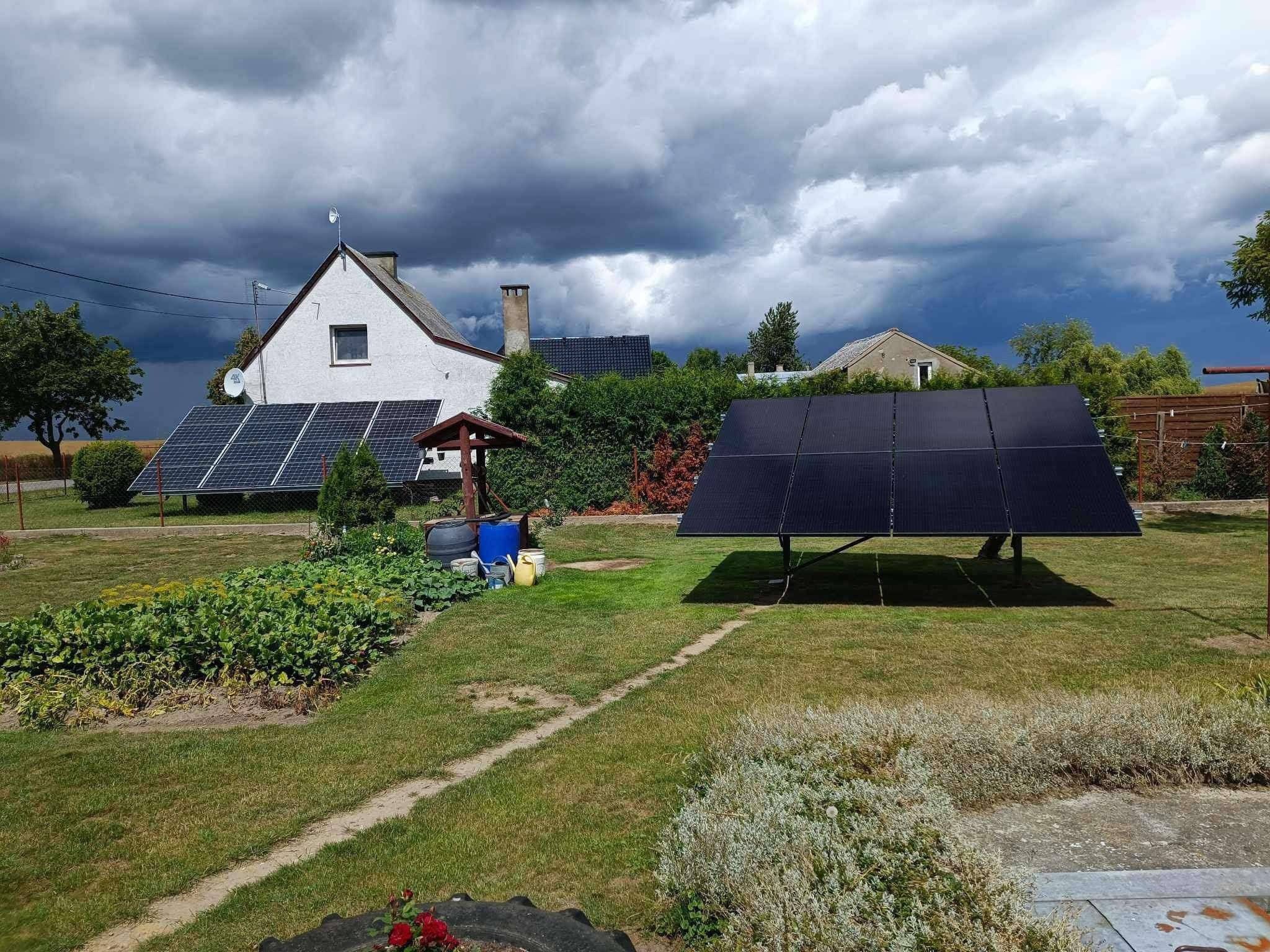 Instalacja fotowoltaiczna na trawniku przed domem z ciemnymi chmurami w tle. Panele ustawione na konstrukcji, widoczne beczki i studnia. Ujęcie w pochmurny dzień.
