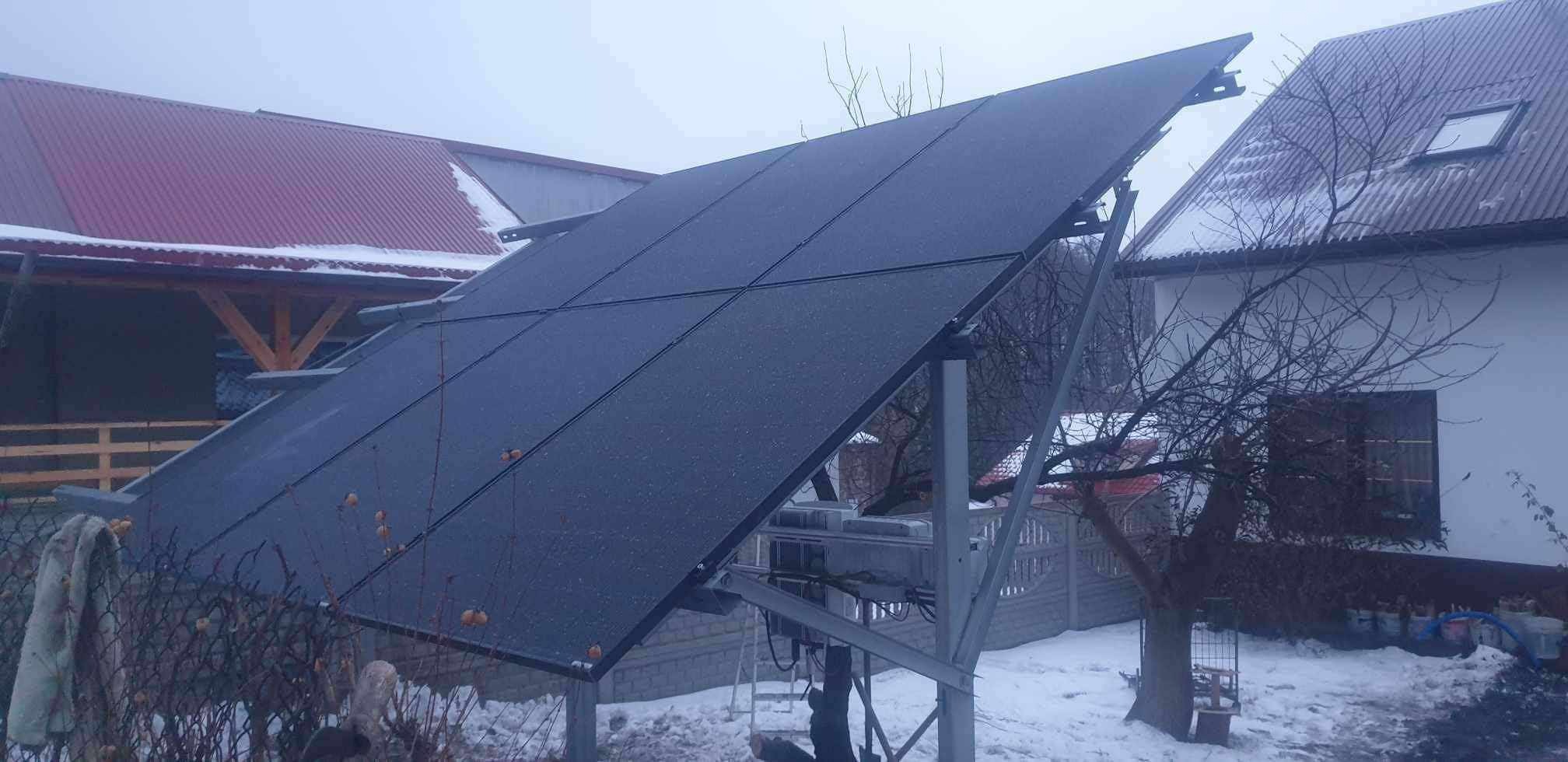 Czarny panel fotowoltaiczny na metalowej konstrukcji, lekko ośnieżony, na tle domu z czerwoną dachówką i zimowego krajobrazu. Widok z dołu, pochmurny dzień.