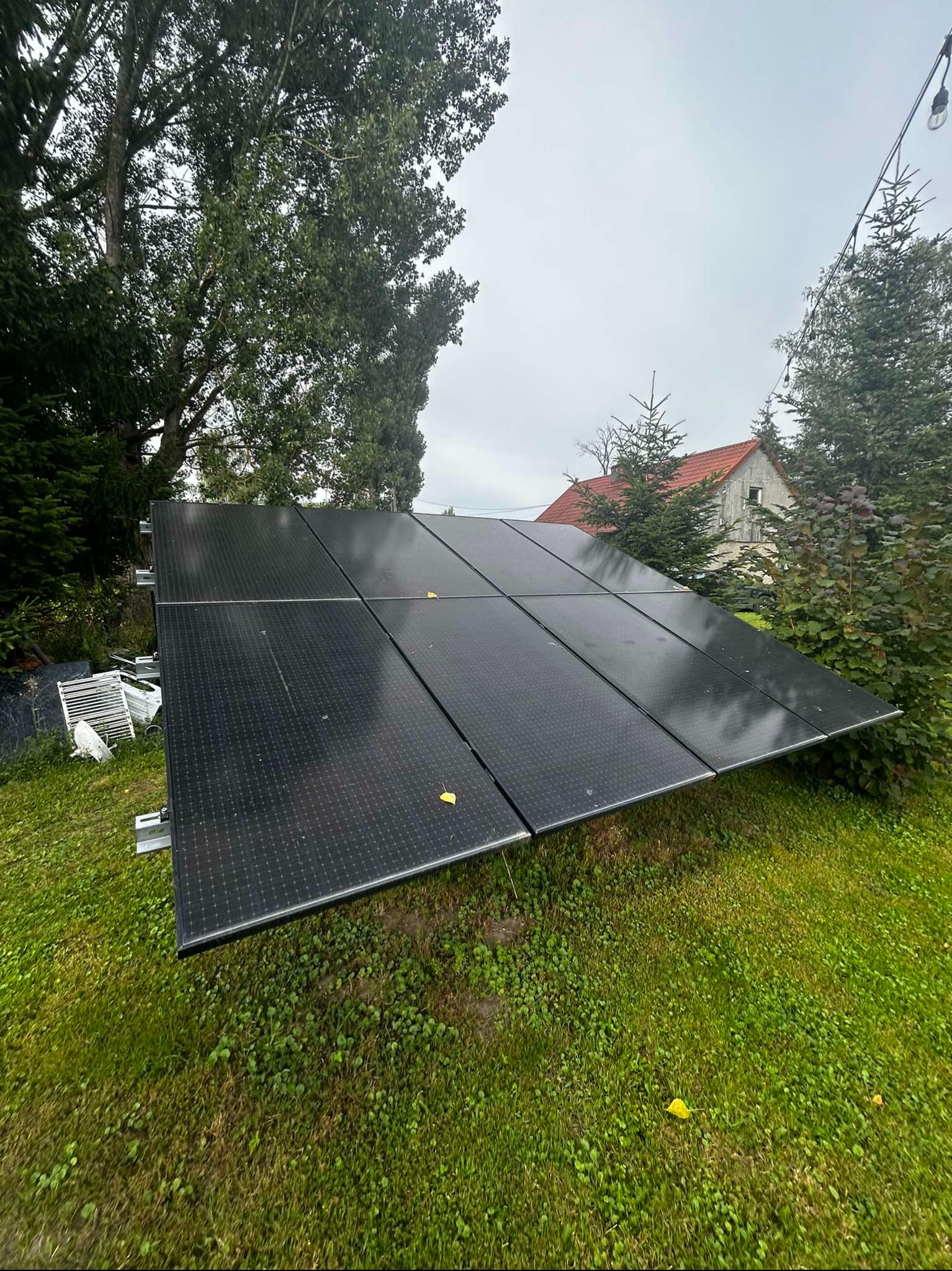 Panel fotowoltaiczny zamontowany na trawie, z widocznym domem w tle i elementami ogrodu. Ujęcie z niskiej perspektywy podkreśla instalację.