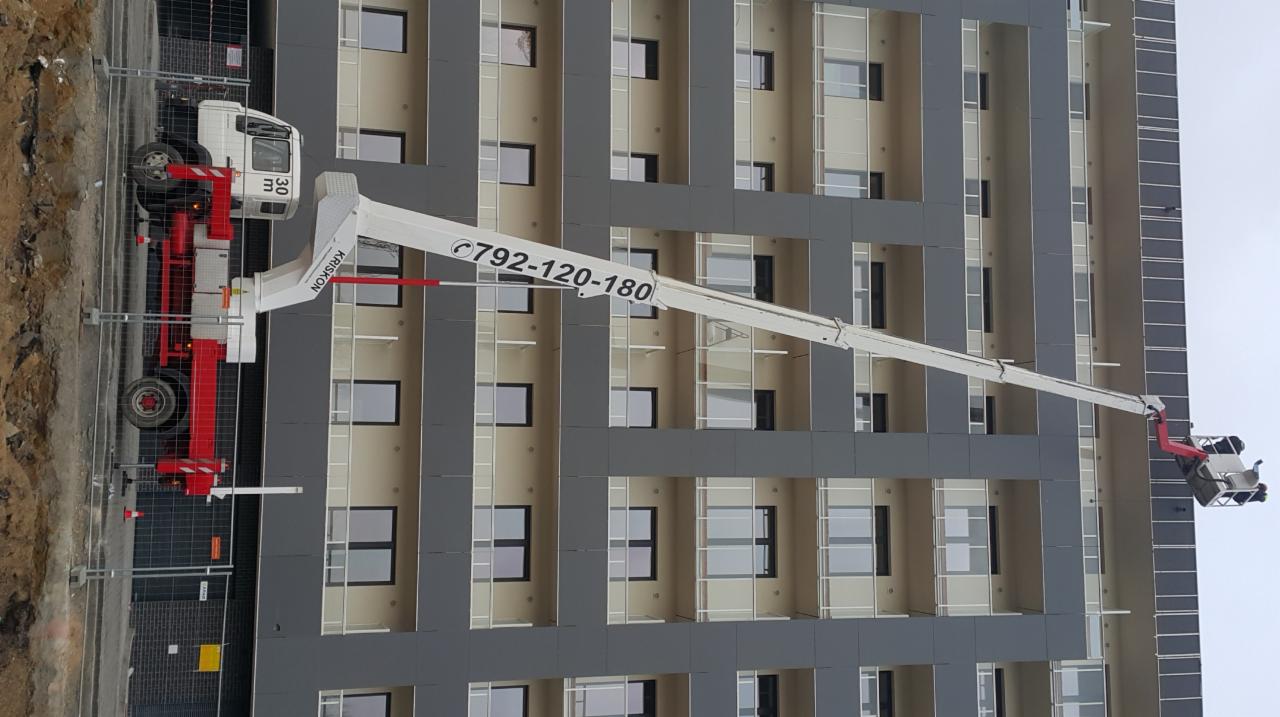Podnośnik koszowy Kriskon z wysięgnikiem sięgającym do balkonu nowoczesnego budynku wielorodzinnego z szarą elewacją, numer telefonu widoczny na ramieniu wysięgnika.