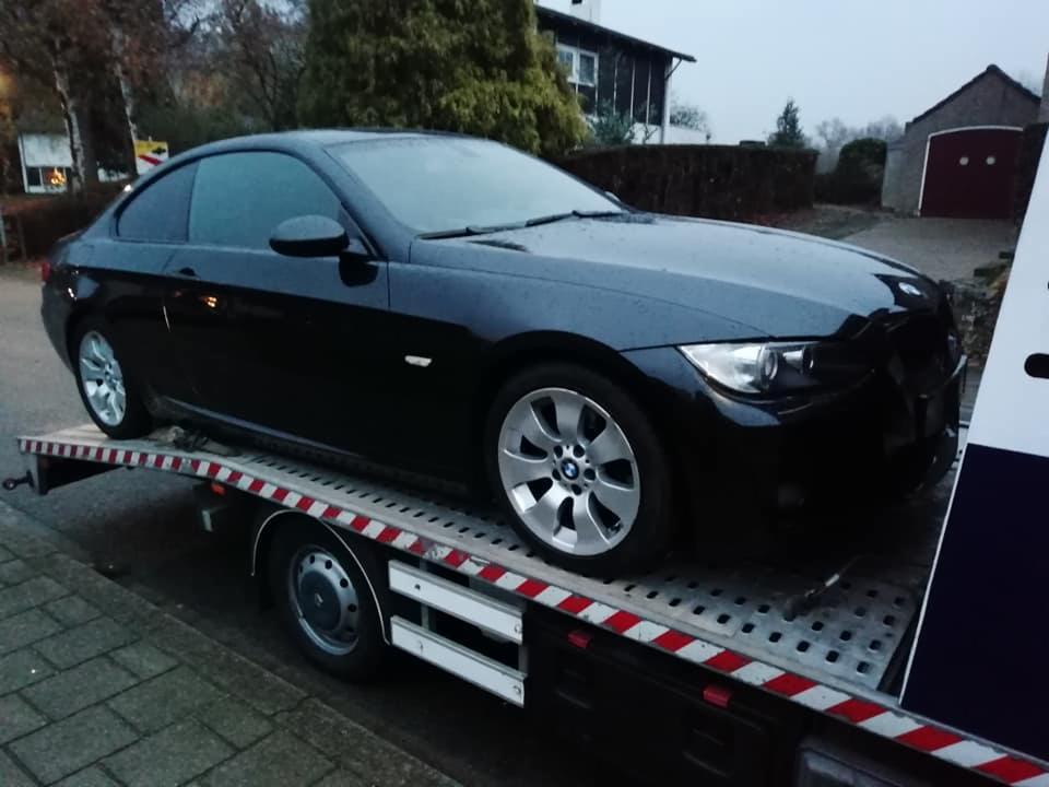 Czarny samochód BMW na platformie lawety z czerwono-białymi pasami, widok z boku na tle domu z ogrodem.
