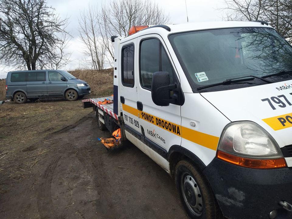 Laweta na błotnistym terenie z częściowo załadowanym szarym busem w tle, widoczny napis 'Pomoc Drogowa' na pojeździe.