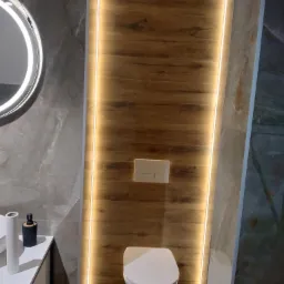 Nowoczesna łazienka z podwieszaną toaletą, drewnopodobnymi panelami ściennymi podświetlonymi listwami LED i okrągłym lustrem z podświetleniem, umywalka z blatem i akcesoriami.