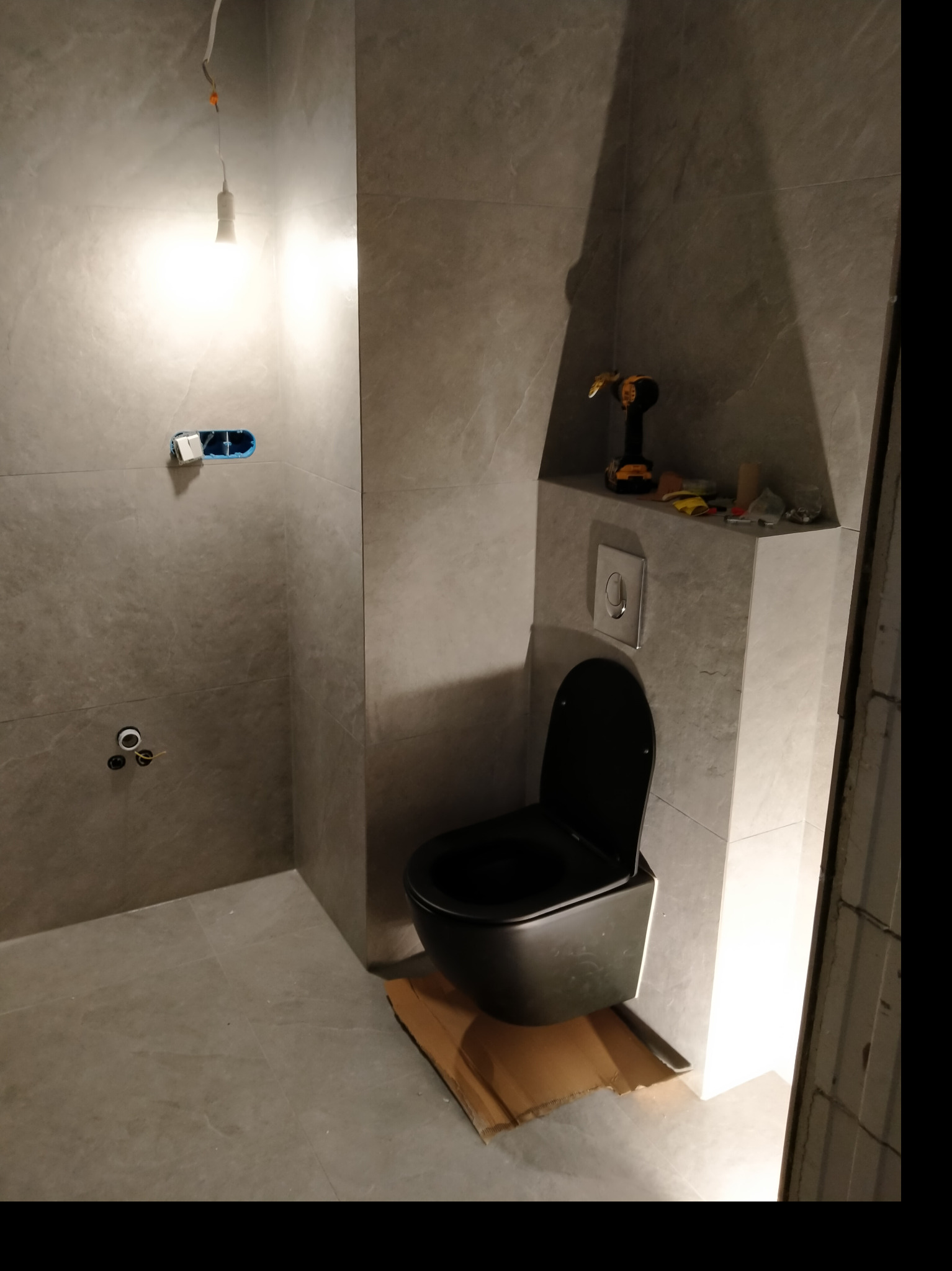 Szara łazienka w trakcie montażu podwieszanego WC, widoczne narzędzia i niezakończone prace elektryczne.