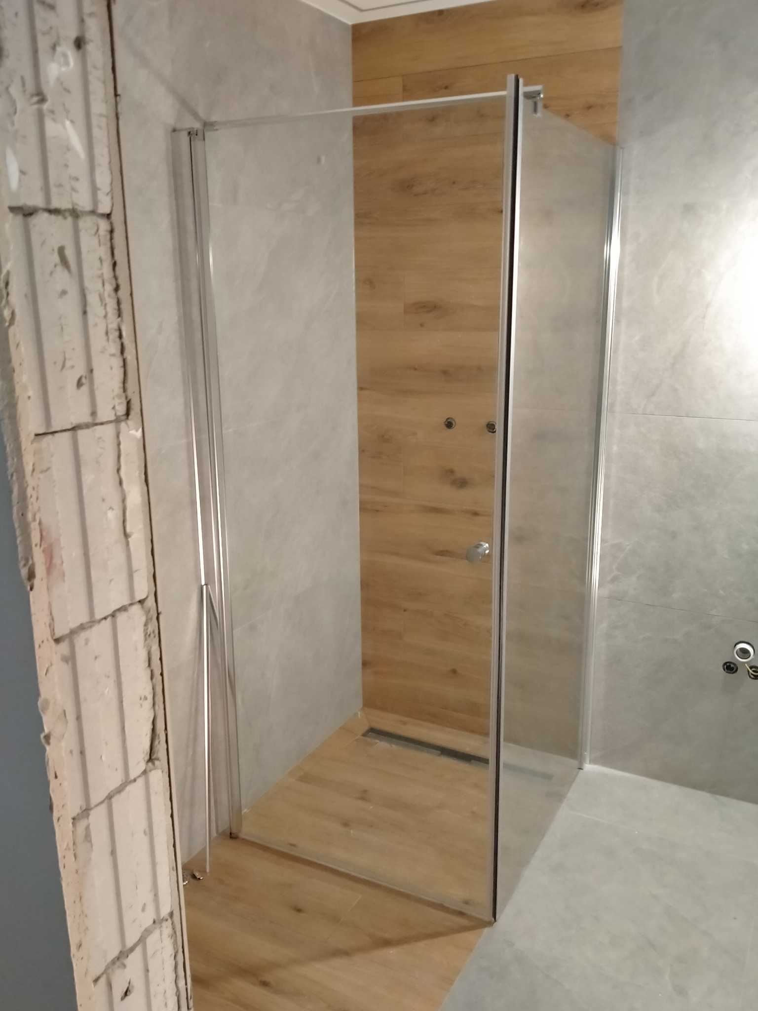 Szklana kabina prysznicowa typu walk-in z odpływem liniowym, zamontowana w łazience z szarymi płytkami i drewnopodobną ścianą, widoczne surowe wykończenie ściany po lewej stronie.