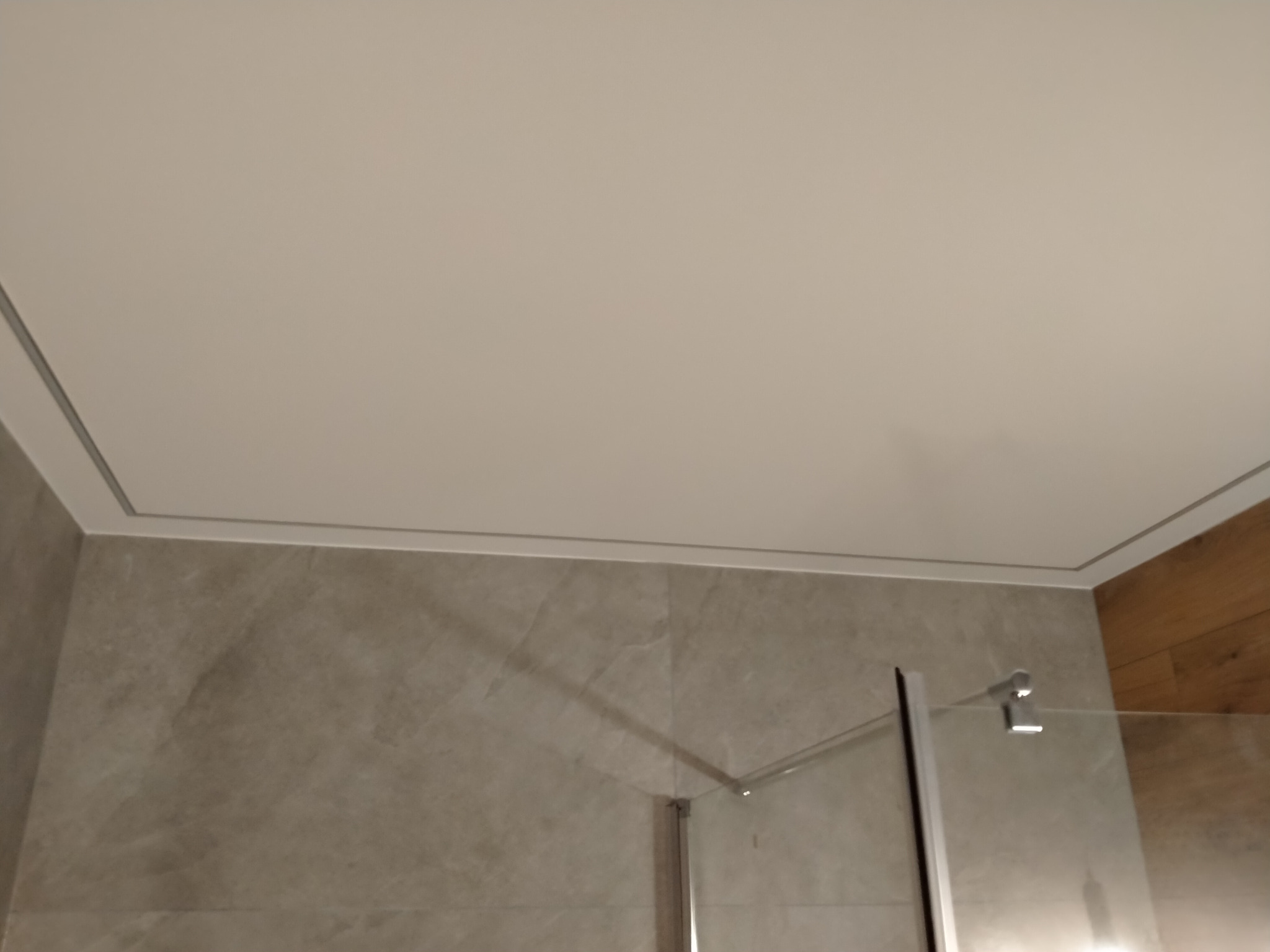 Wykończona łazienka: gładki, beżowy sufit podwieszany z delikatną listwą ozdobną, szare płytki imitujące kamień na ścianie, częściowo widoczna szklana kabina prysznicowa z chromowanymi elementami...