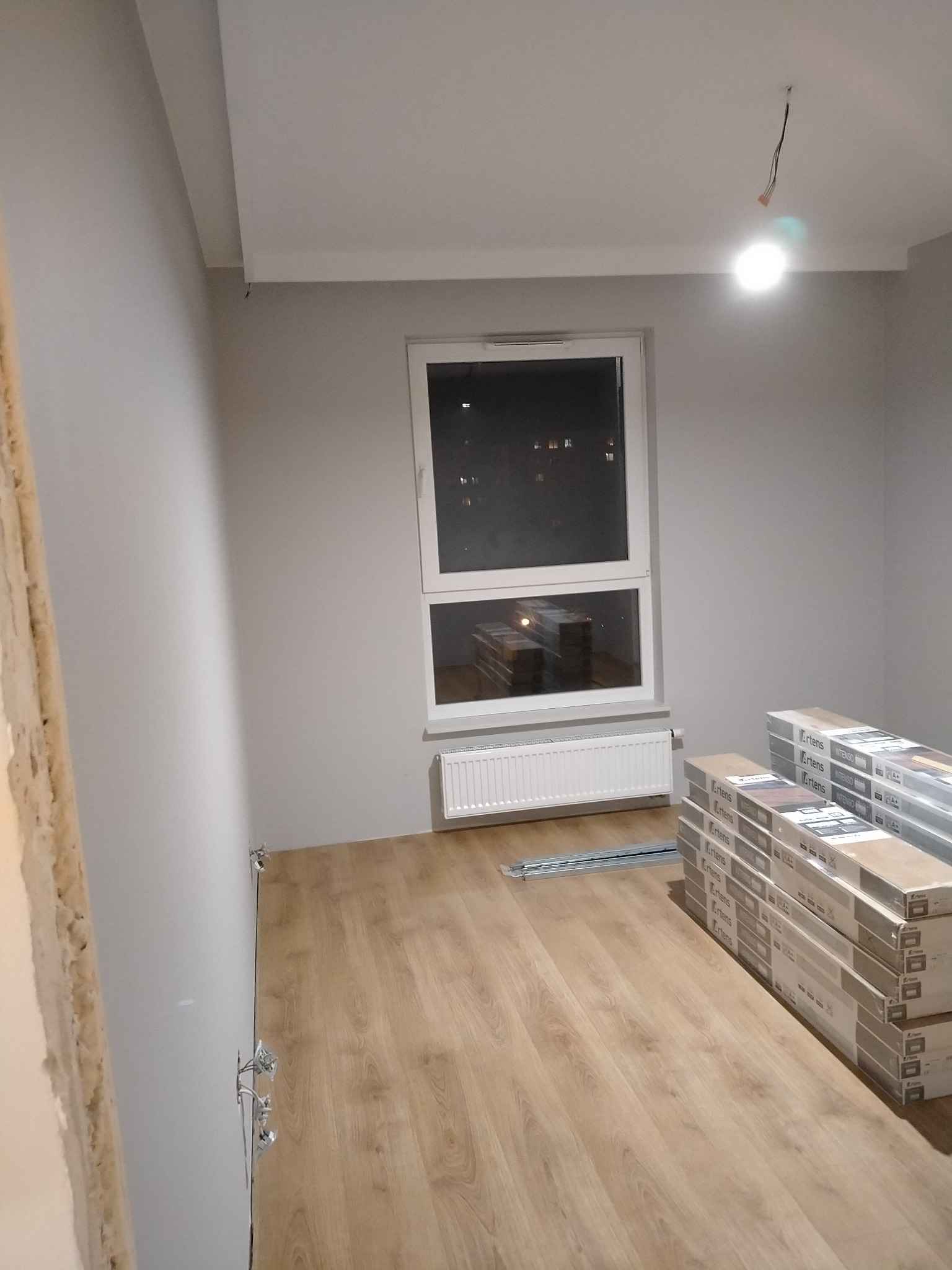 Pokój w trakcie układania paneli podłogowych, widoczne paczki paneli marki Artens, grzejnik pod oknem, surowe wykończenie ścian, wystające kable elektryczne z sufitu i gniazdek.
