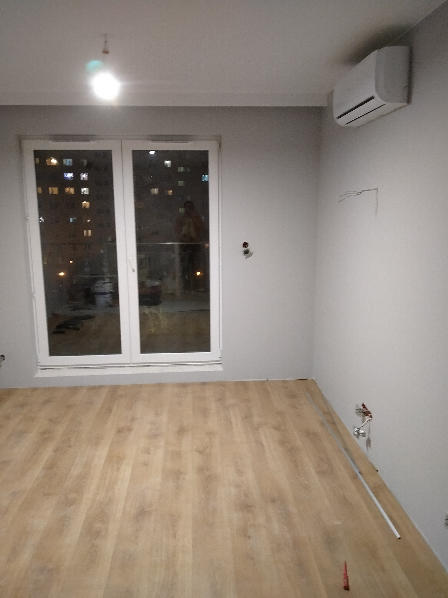 Pusty pokój po remoncie z nową podłogą z paneli, klimatyzatorem na ścianie i widocznymi instalacjami elektrycznymi. W oknach odbija się nocny widok miasta.