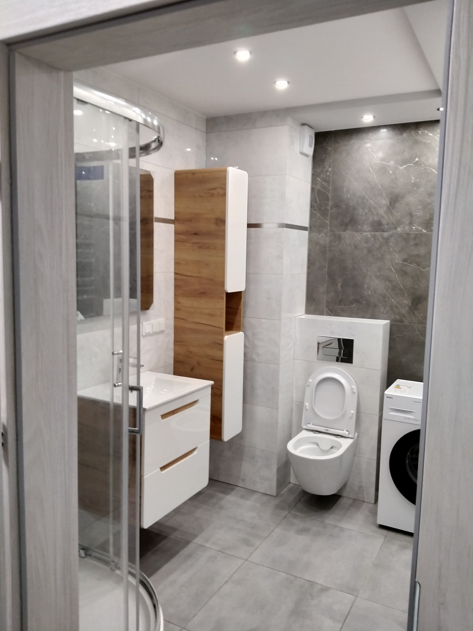 Nowoczesna łazienka w Turce z kabiną prysznicową, umywalką z szafką, toaletą podwieszaną, pralką i płytkami imitującymi marmur na ścianie.