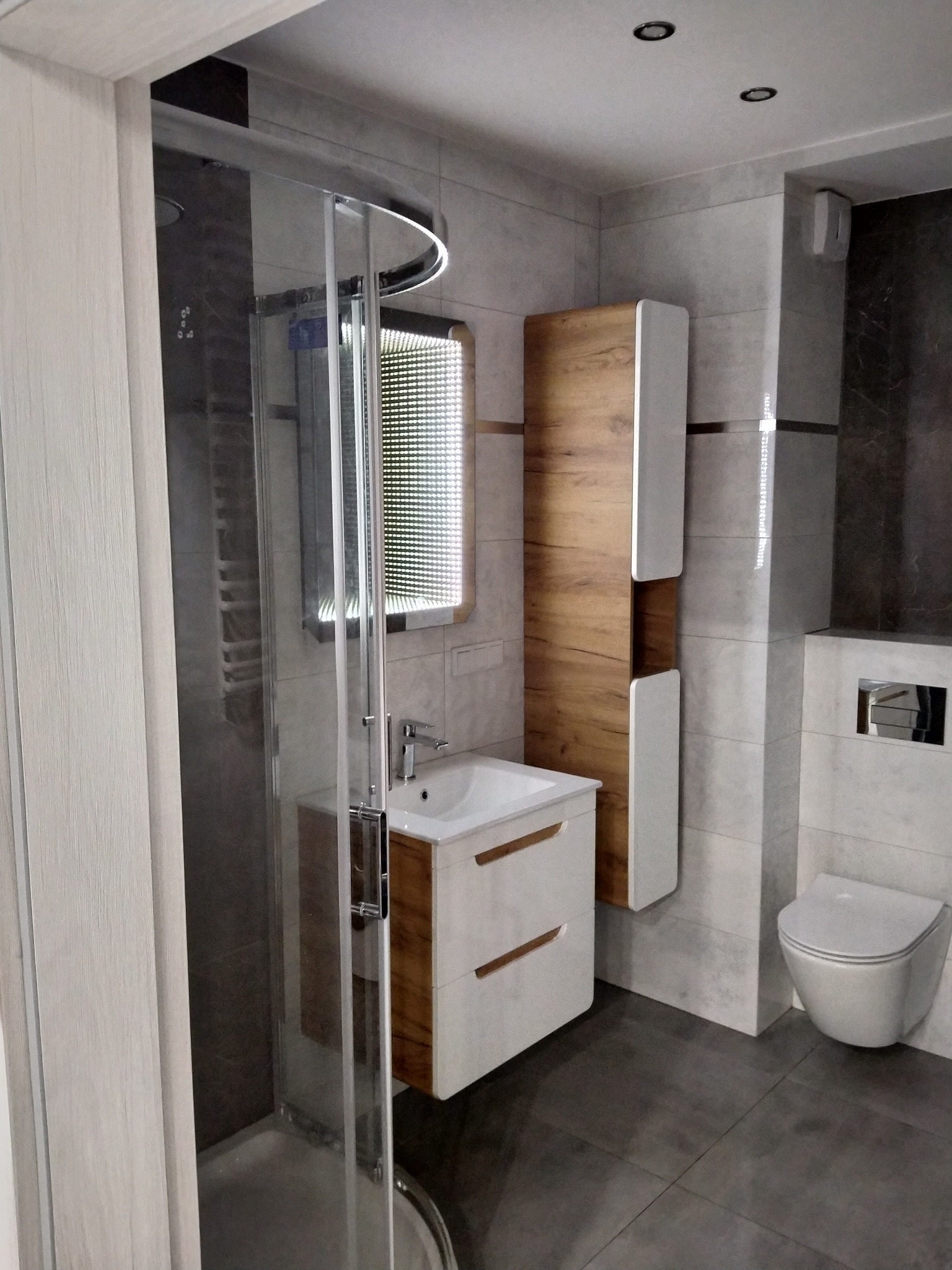 Nowoczesna łazienka z kabiną prysznicową z zaokrąglonymi drzwiami, umywalką z szafką w odcieniach bieli i drewna, wiszącą toaletą oraz szarymi płytkami na ścianach i podłodze.