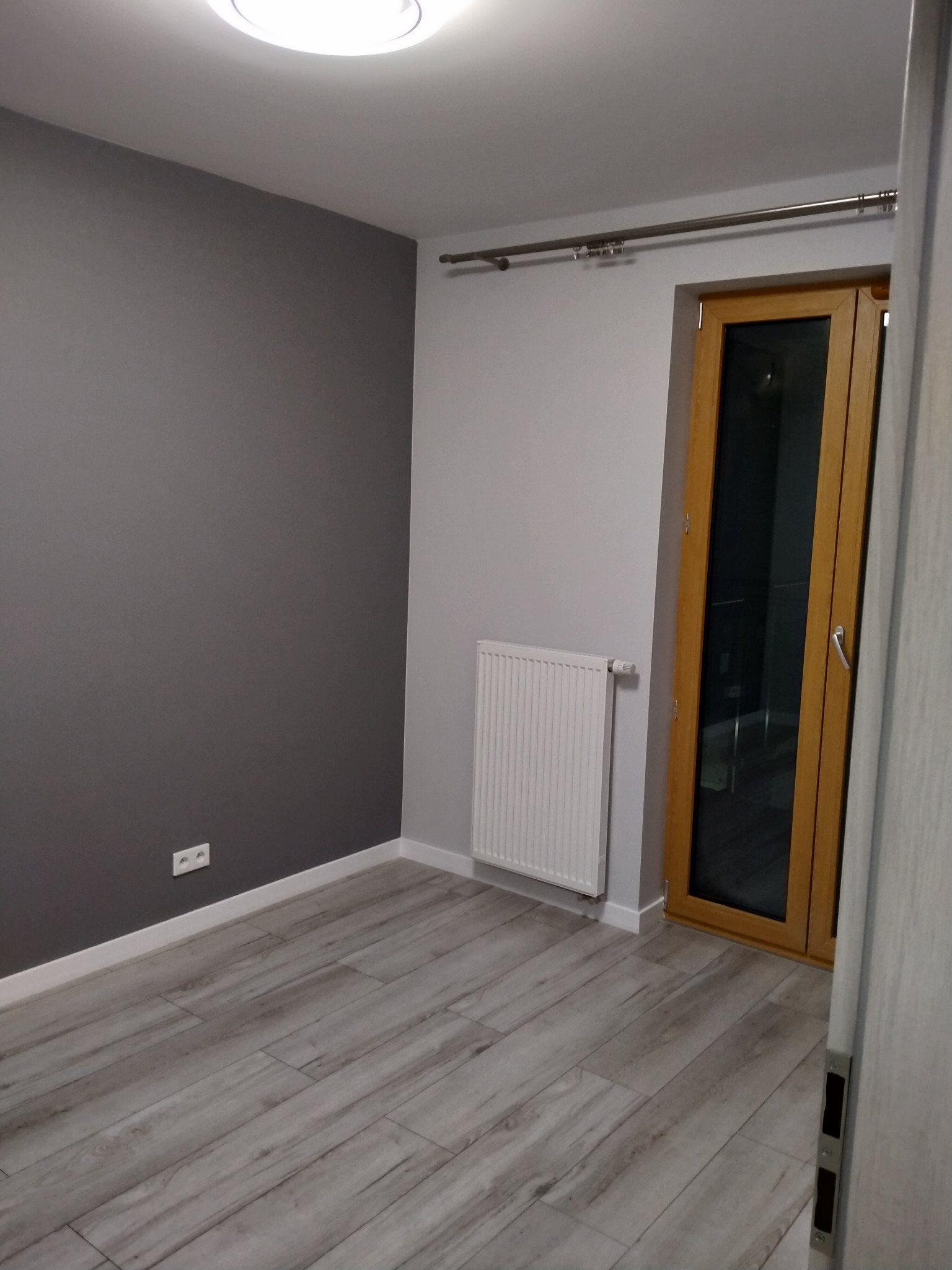Pokój po odświeżeniu: szara ściana, jasne panele podłogowe, biały grzejnik i drewniane drzwi balkonowe z metalowym karniszem.
