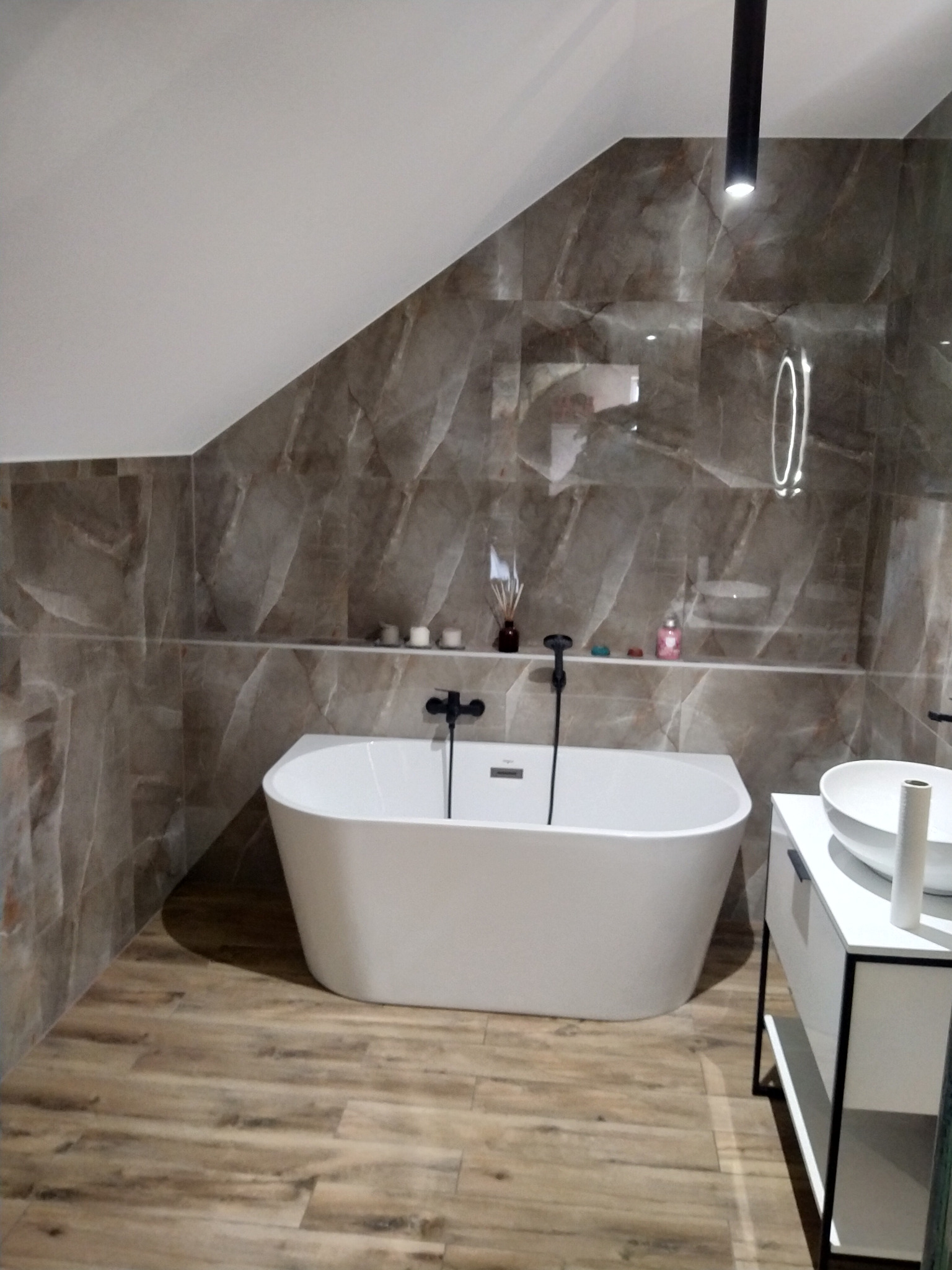 Nowoczesna łazienka na poddaszu z wolnostojącą białą wanną, marmurowymi płytkami na ścianach i drewnopodobną podłogą, oświetlona punktowo czarnym reflektorem.