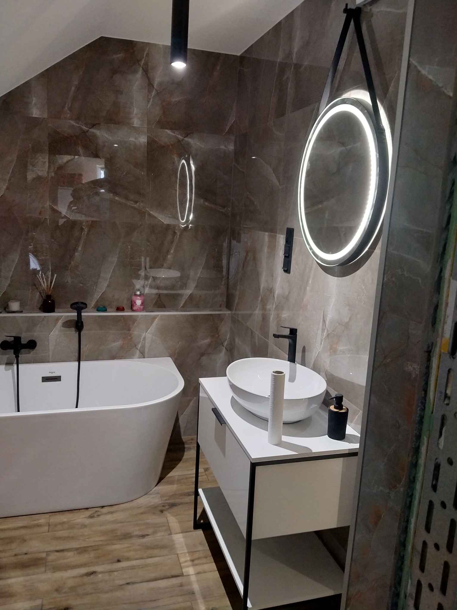 Nowoczesna łazienka na poddaszu z brązowymi płytkami imitującymi marmur, okrągłe lustro LED na pasku, biała wolnostojąca wanna i umywalka na szafce z czarnymi elementami
