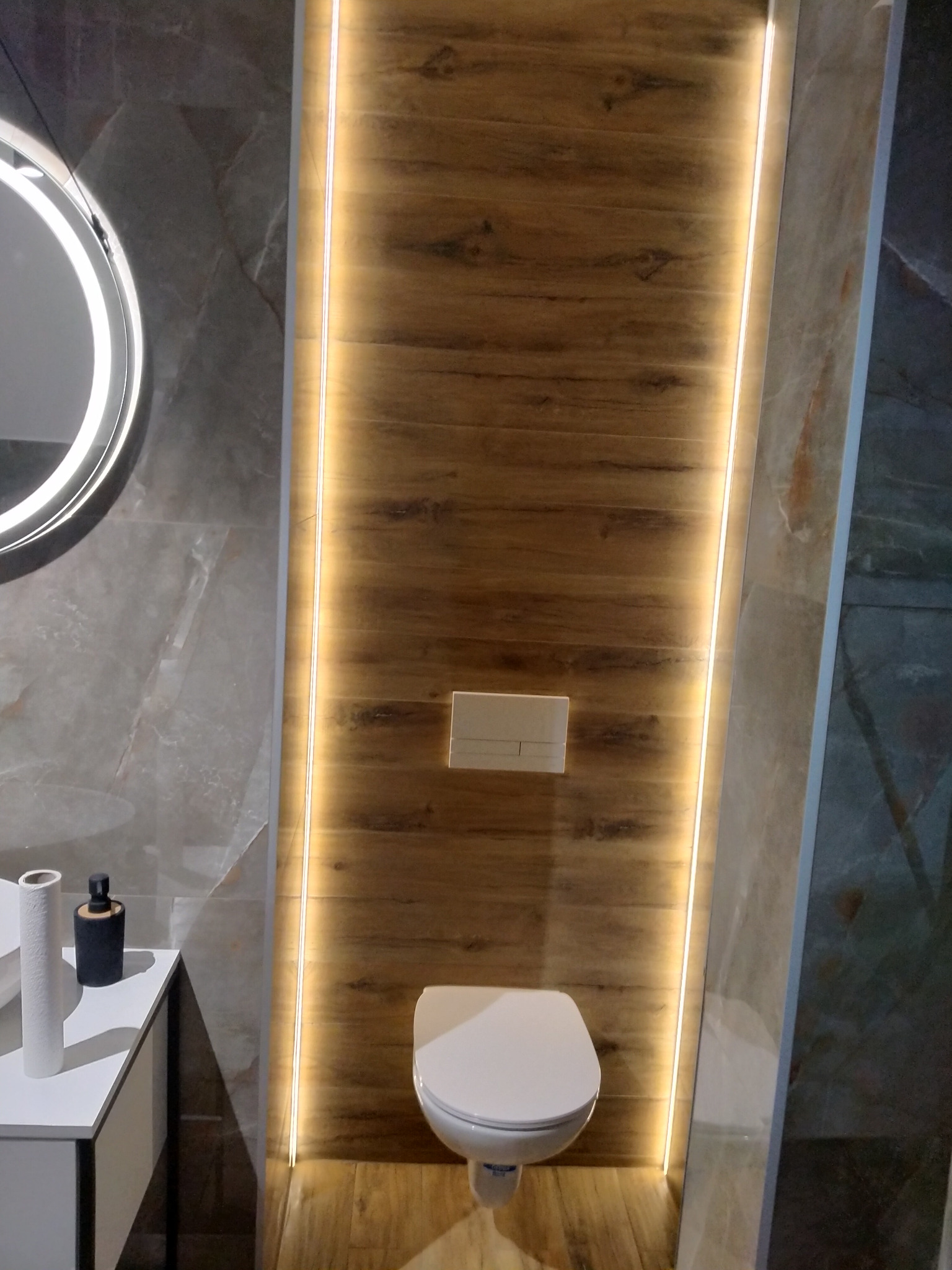 Nowoczesna łazienka z podwieszaną toaletą, drewnopodobnymi panelami ściennymi podświetlonymi listwami LED i okrągłym lustrem z podświetleniem, umywalka z blatem i akcesoriami.