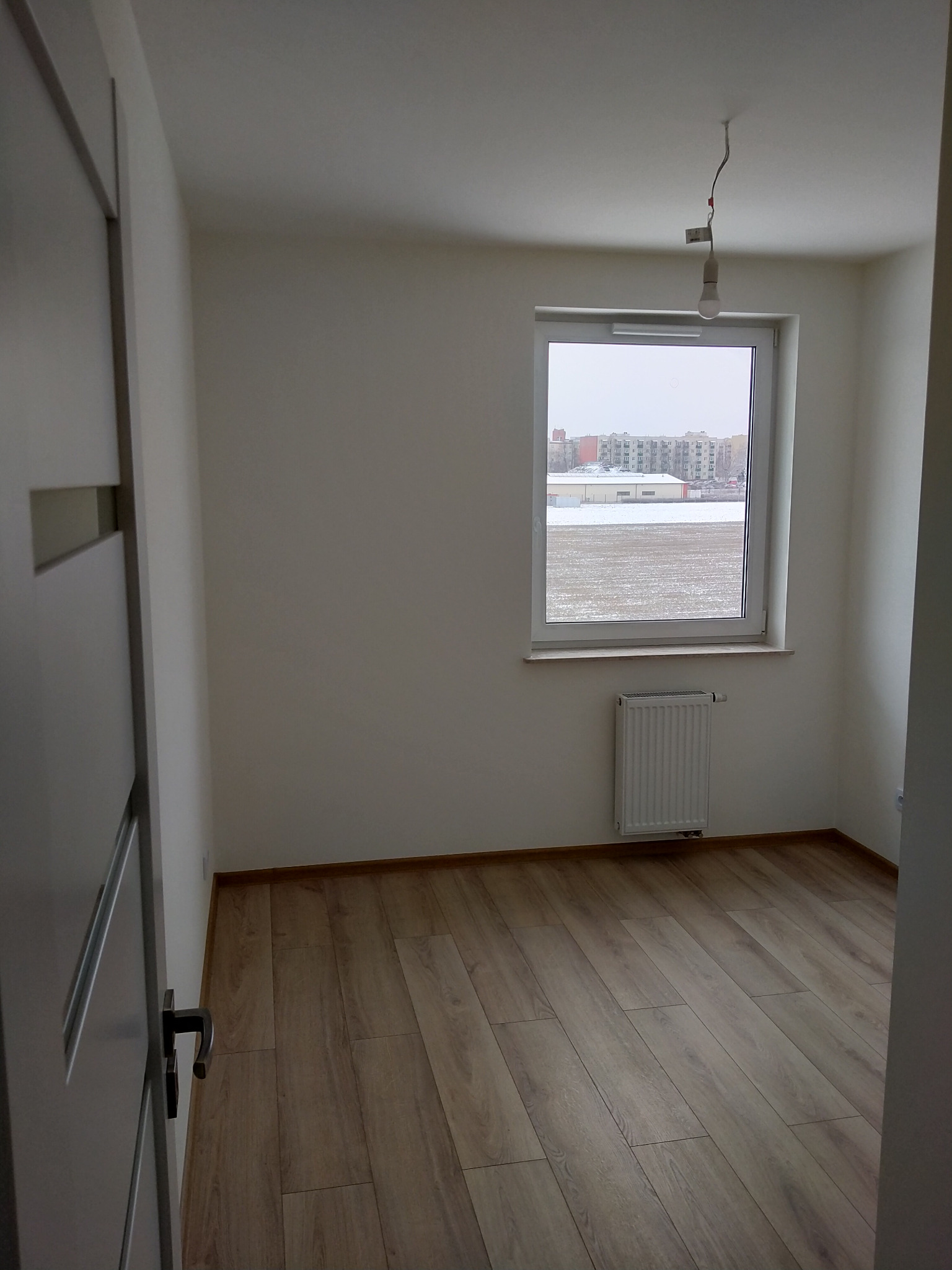 Pusty pokój z panelami podłogowymi w odcieniach brązu, białymi ścianami, grzejnikiem pod oknem z widokiem na zimowy krajobraz i niedokończoną instalacją oświetleniową.