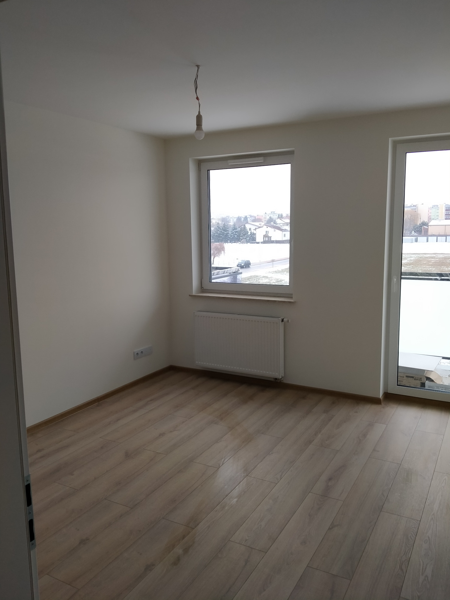 Pusty pokój z panelami podłogowymi w odcieniach brązu, świeżo pomalowanymi ścianami na biało, grzejnikiem pod oknem i wiszącą żarówką bez klosza.