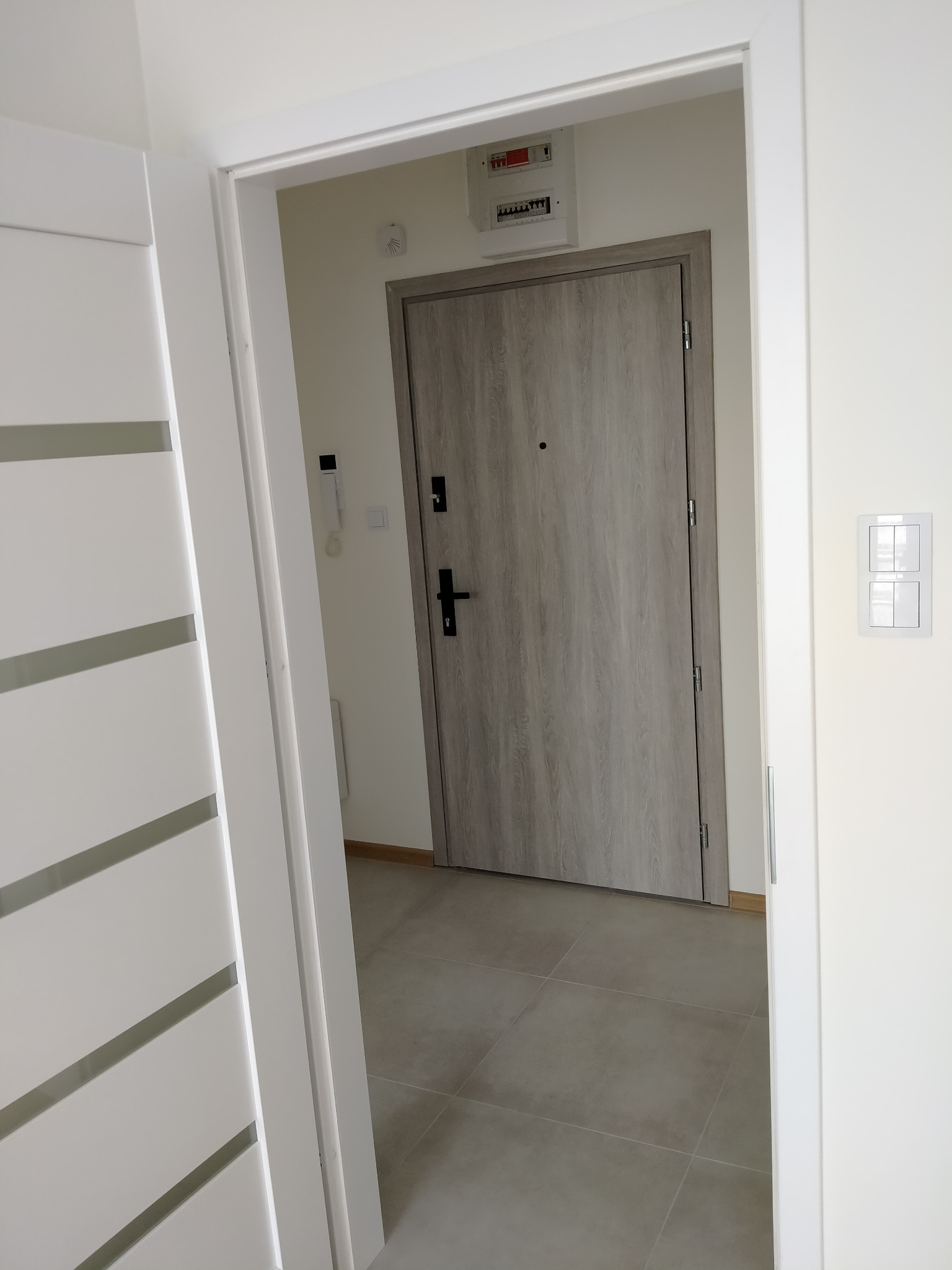 Otwarta biała, nowoczesna drzwi wewnętrzne z poziomymi przeszkleniami, widok na szarą, drewnopodobną drzwi wejściowe do mieszkania i szarą podłogę z płytek